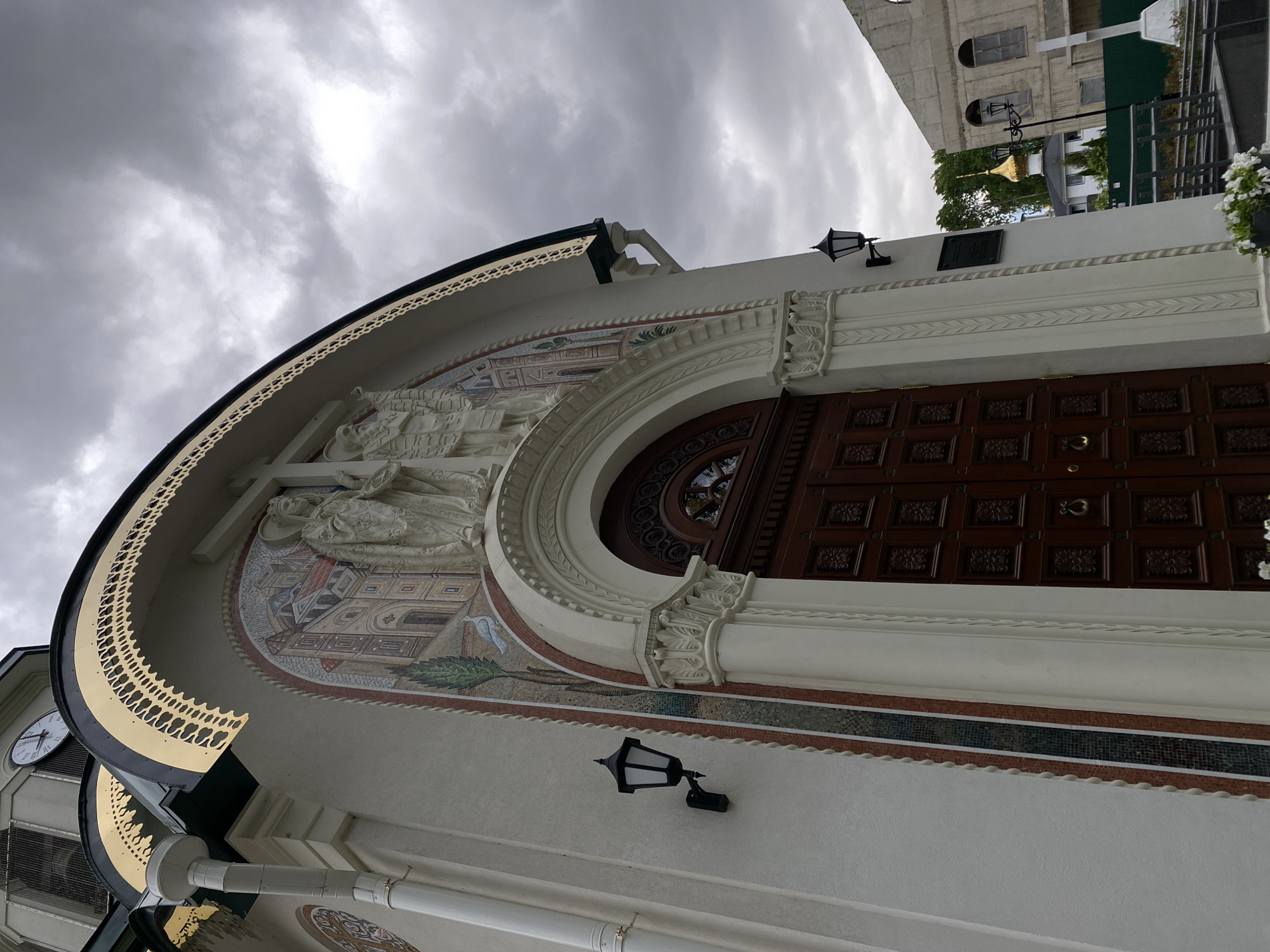 This image depicts the facade of the Church of the Savior on the Spilled Blood, located in Saint Petersburg, Russia. The facade features a large arched entrance with intricate mosaic artwork depicting religious figures and scenes. Above the arch, there are sculptural elements. The exterior wall is white with decorative trim and a dark wooden door. A decorative lamp is mounted on the wall, and elements of a clock tower are visible to the left. The sky is overcast.
