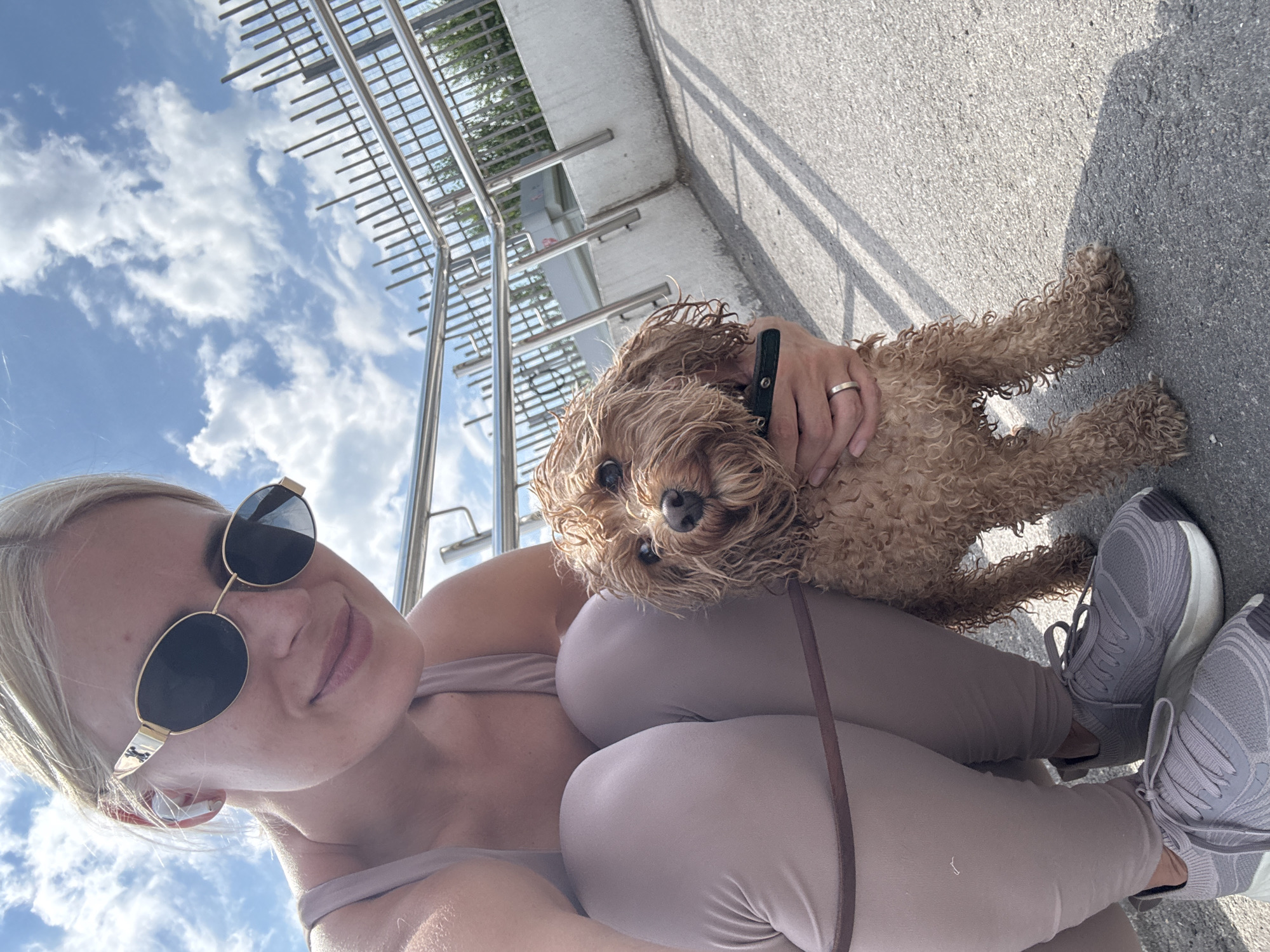 A blonde woman wearing sunglasses and athletic wear is crouching on a paved surface, holding a small, curly-haired dog. The woman has earbuds in her ears and is wearing a ring on her left hand. The dog is brown and appears to be wet. Behind them is a metal railing and a concrete structure. The sky is blue with white clouds. The scene appears to be outdoors, possibly in a park or public walkway. There is no indication that this location is in Saratov, Russia.