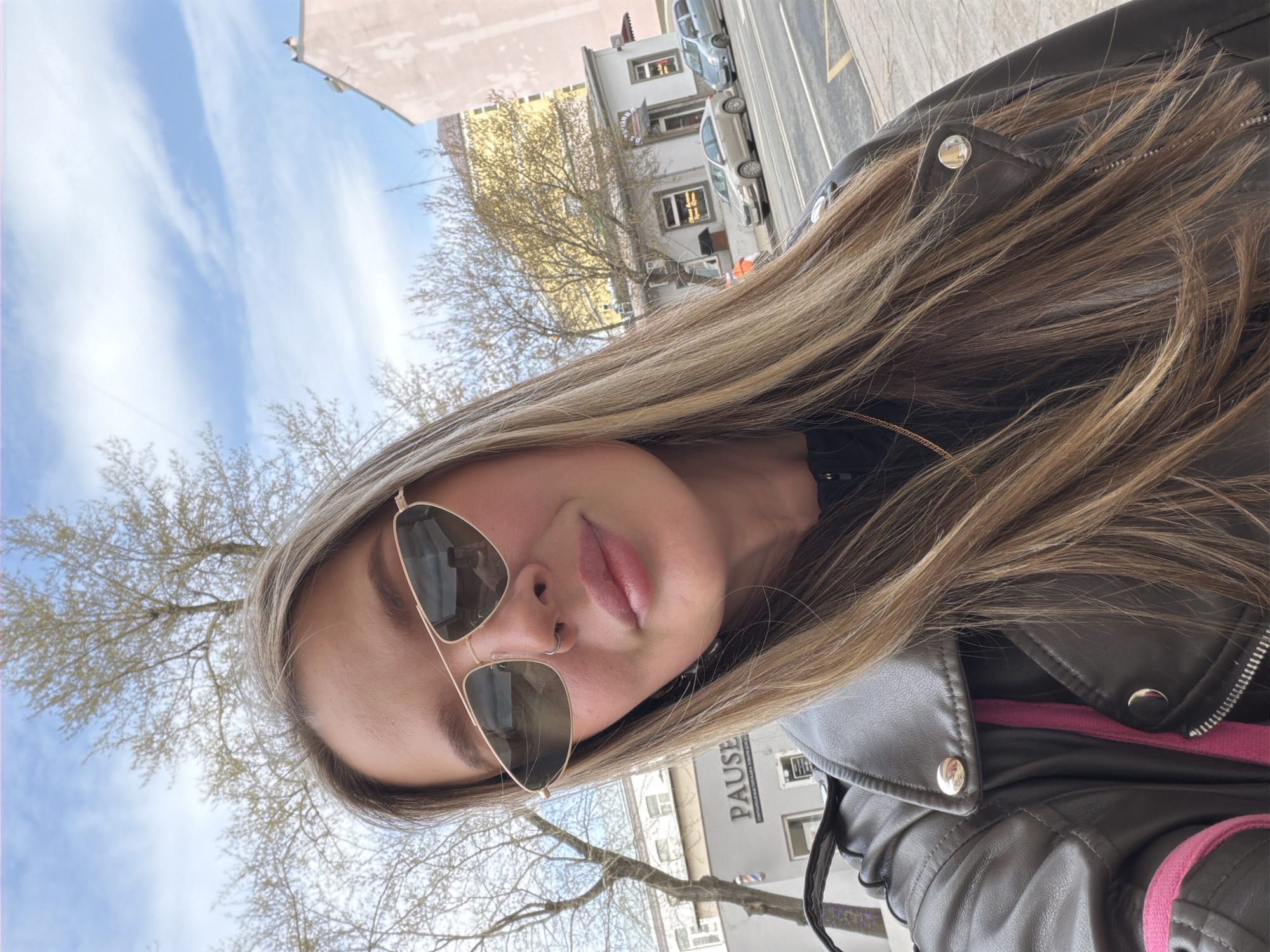A young woman poses for a selfie outdoors on a sunny day in Moscow, Russia. She wears aviator sunglasses, a black leather jacket over a pink top, and has long blonde hair. The background includes buildings, trees with budding leaves, a blue sky with scattered clouds, and parked cars on a street. The word "PAUSE" is visible on one of the buildings.