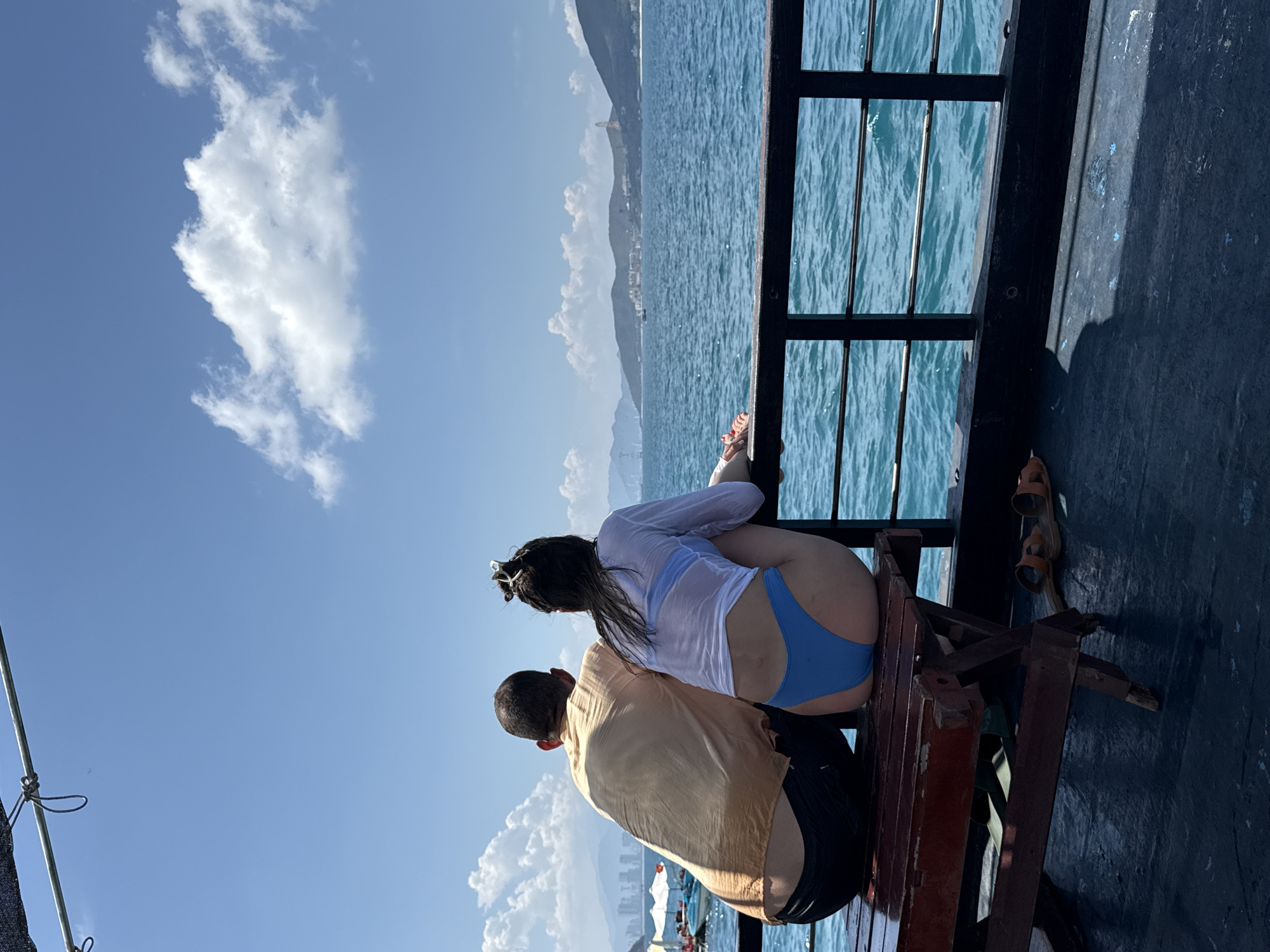 A couple is sitting on a bench facing the sea, likely on a boat. The woman is wearing a white top and blue swimsuit bottoms. The man is wearing a tan shirt and black shorts. The sea is blue with some waves. In the background, there are mountains and some buildings which suggest the location is Nam Nha Trang Ward, Vietnam. There is a cloudy blue sky above. The bench appears to be on a boat or a dock with a railing.