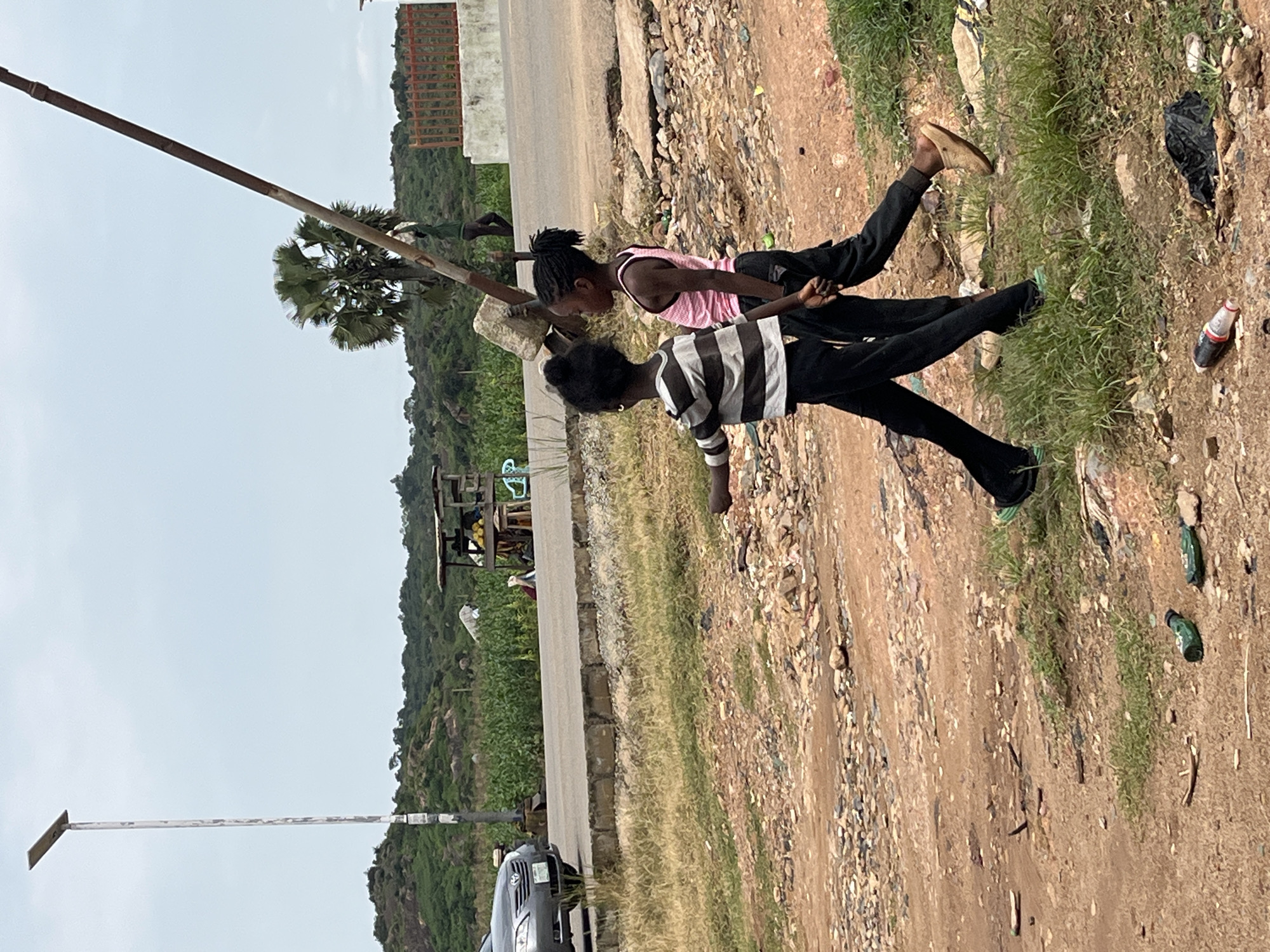 Two young girls, one wearing a pink tank top and black pants and the other a striped shirt and black pants, walk past a tilted pole in a rural area. The landscape is characterized by a dirt road, overgrown grass, and rocky terrain. In the background, a car drives on a paved road, and a small roadside stall selling produce is visible. Further in the distance, a wooded area and hills under a cloudy sky complete the scene. The image was captured on a street in Jos, Nigeria.