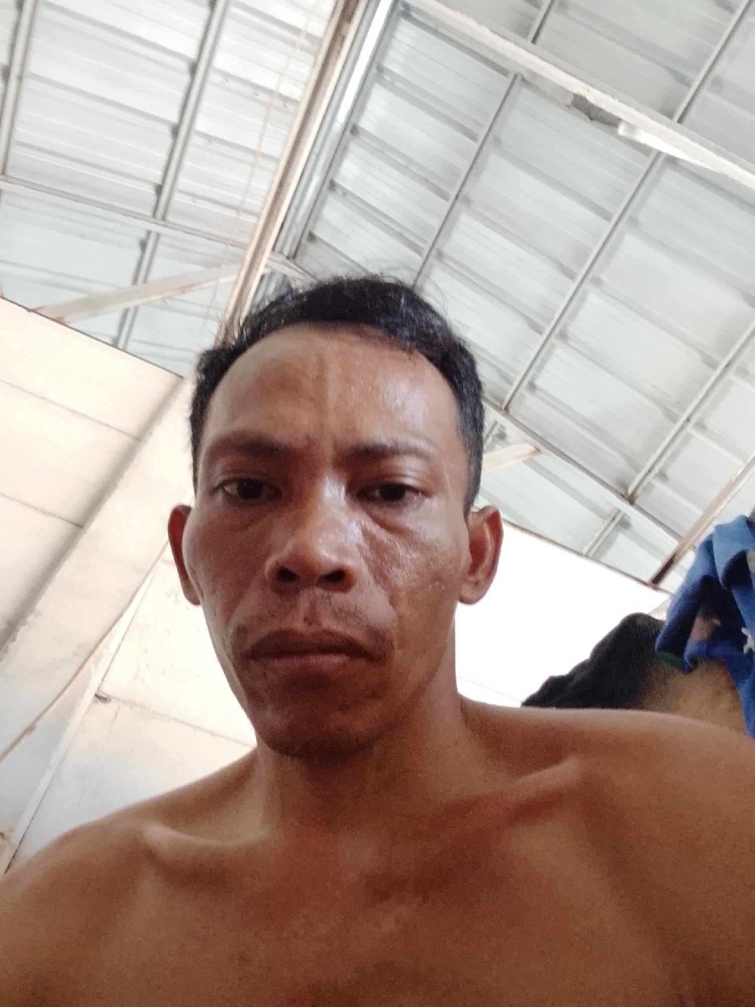 The image shows a close-up, low-angle selfie of a middle-aged man with short, dark hair. He has a serious expression. His skin appears weathered. He is shirtless. The background is a corrugated metal ceiling, suggesting an interior location. The image was apparently taken in Bukit Ketok, Indonesia. A partially visible blue cloth is present behind his right shoulder.
