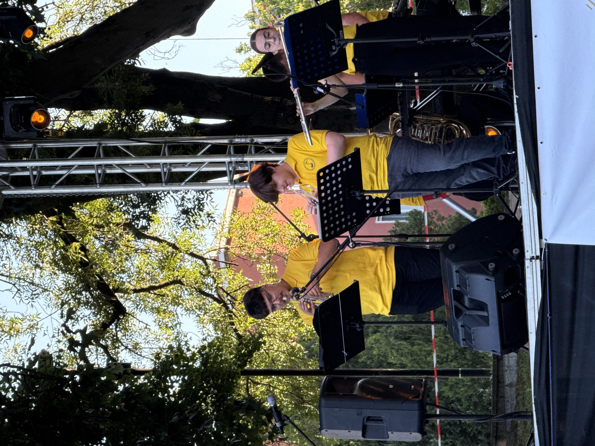 An outdoor concert is taking place on a stage. Four young people, presumably members of a band, are playing instruments. Two individuals in the foreground are wearing yellow polo shirts and dark pants. One is playing a trumpet, and the other is playing a flute. Behind them, two other individuals are also playing instruments. The stage is set up in front of trees and a building. The lighting appears to be natural, with some stage lights visible in the background. The overall atmosphere suggests a performance or rehearsal during the daytime. The location is specified as the city of Pleven, Bulgaria.
