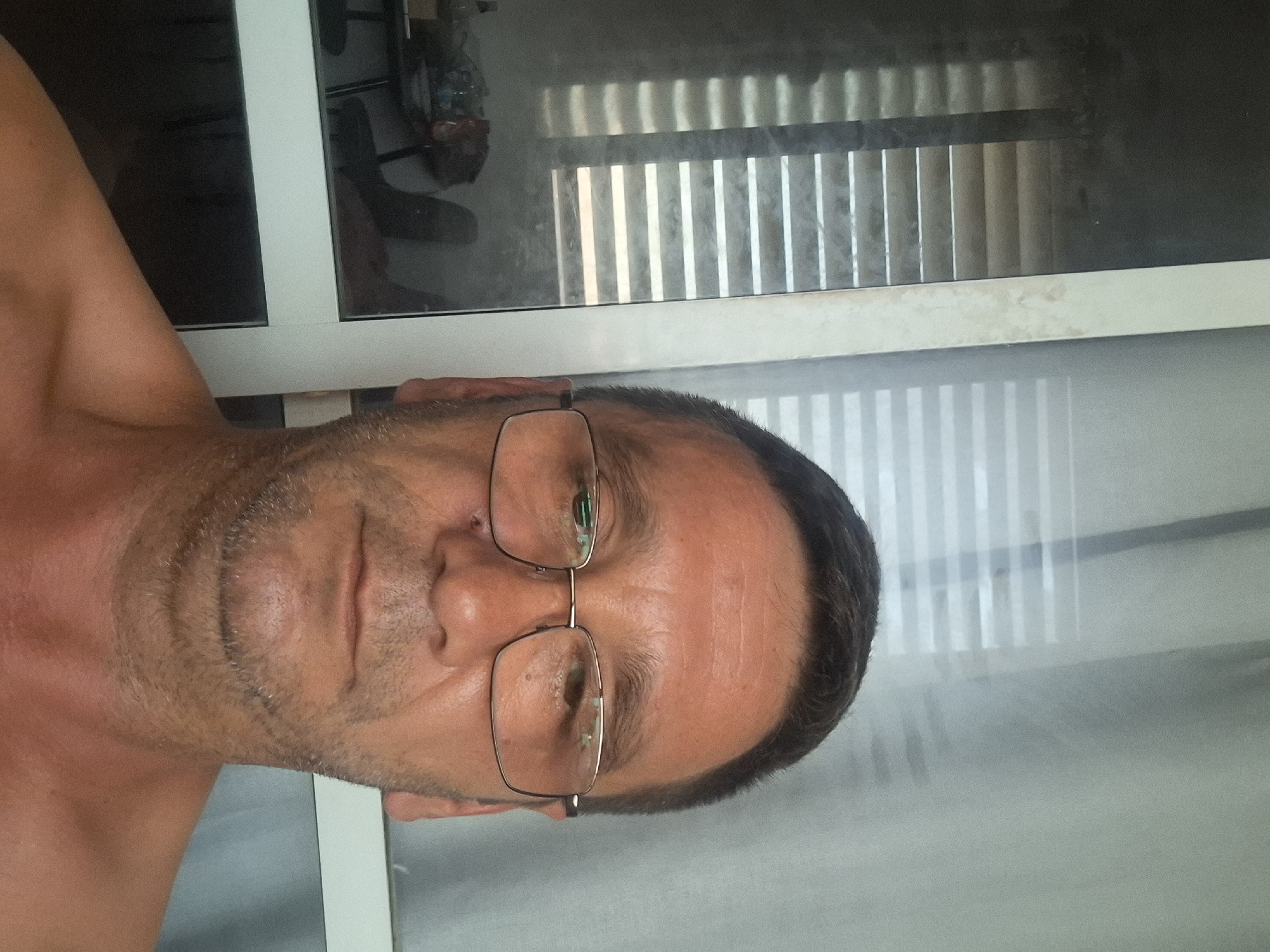 The image shows a close-up, shoulder-up, selfie-style photograph of a man with short, dark hair, wearing glasses. He has stubble on his face and a small mark or blemish near his left eye. The background consists of sheer, light-colored curtains with horizontal blinds visible through them. To the left of the man, a reflection in glass reveals a room with furniture and what appears to be a table with items on it. The overall lighting suggests an indoor setting, possibly during the day. The location is identified as Yerocham, Israel.