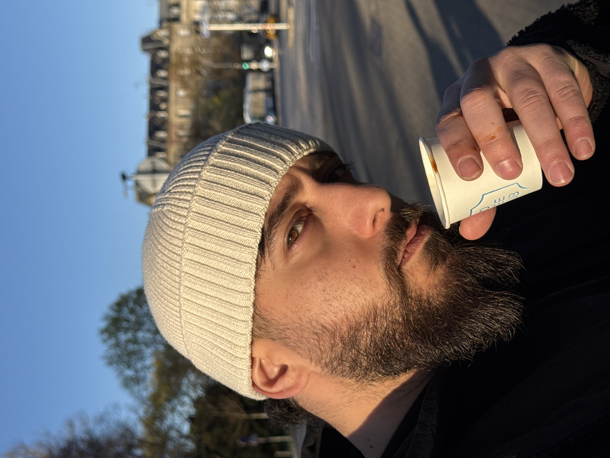 An outdoor, close-up portrait shows a male subject in Strasbourg, France, holding a white paper cup. The man possesses a full, dark beard and mustache, brown eyes, and wears an off-white ribbed knit beanie. He is clad in a dark-colored, possibly black, textured jacket. Strong, directional sunlight illuminates the right side of his face (viewer's left), casting warm light and distinct shadows. His left hand, holding a small white paper cup with a partial blue logo, shows minor reddish-brown stains on the fingers and cup rim. The background is out of focus but reveals an urban street scene under a clear blue sky, featuring multi-story buildings with classic architecture, a wide paved road, a blurred white public transport vehicle (bus or tram), a visible green traffic light, and trees with some bare branches, suggesting an avenue or park setting adjacent to the street.