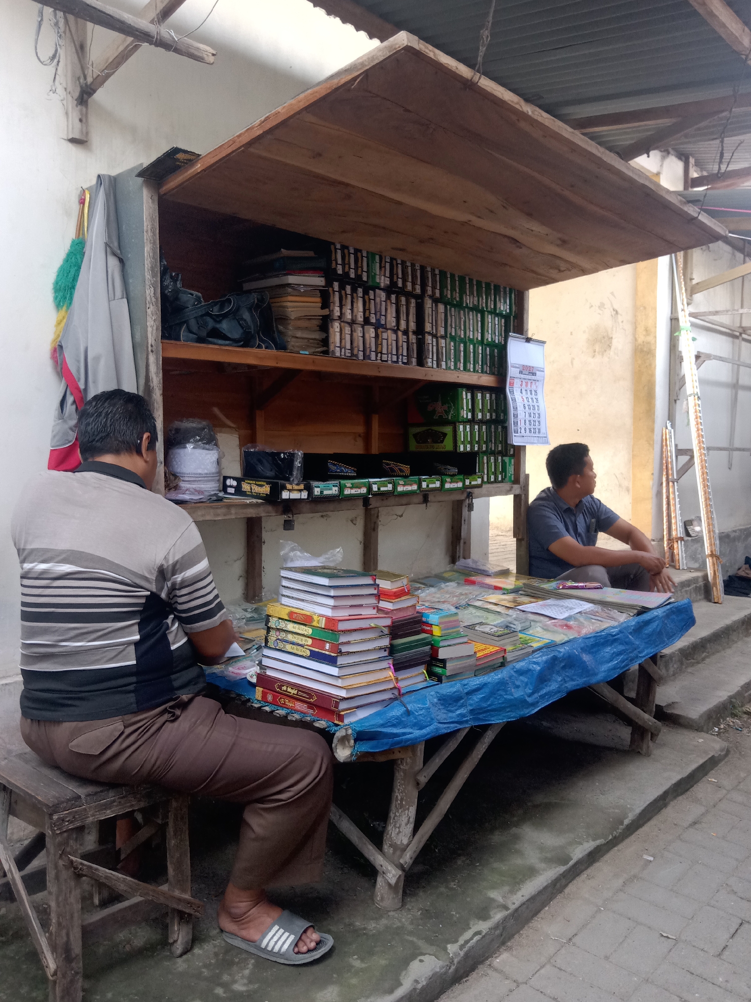 In the city of Klepek, Indonesia, a street vendor is shown operating a small stall under a wooden awning. Two men are present at the stall. One man, wearing a striped t-shirt and brown pants, sits on a wooden stool, facing away from the camera and towards the stall. The other man, in a blue t-shirt and grey pants, sits on the ground to the right of the stall, facing left and looking away from the camera.

The stall is laden with goods, primarily books stacked in piles on a blue tarp-covered table. Behind the books, on shelves, are rows of what appear to be small boxes or containers, possibly religious texts or related items, and some larger books. A calendar for 2021 is hanging on the back wall of the stall. Several items, including what looks like prayer caps and bags, are also visible on the shelves. The stall itself is constructed from wood and appears weathered. The ground around the stall is paved with concrete and some cobblestone. The environment suggests an outdoor market or a side street.