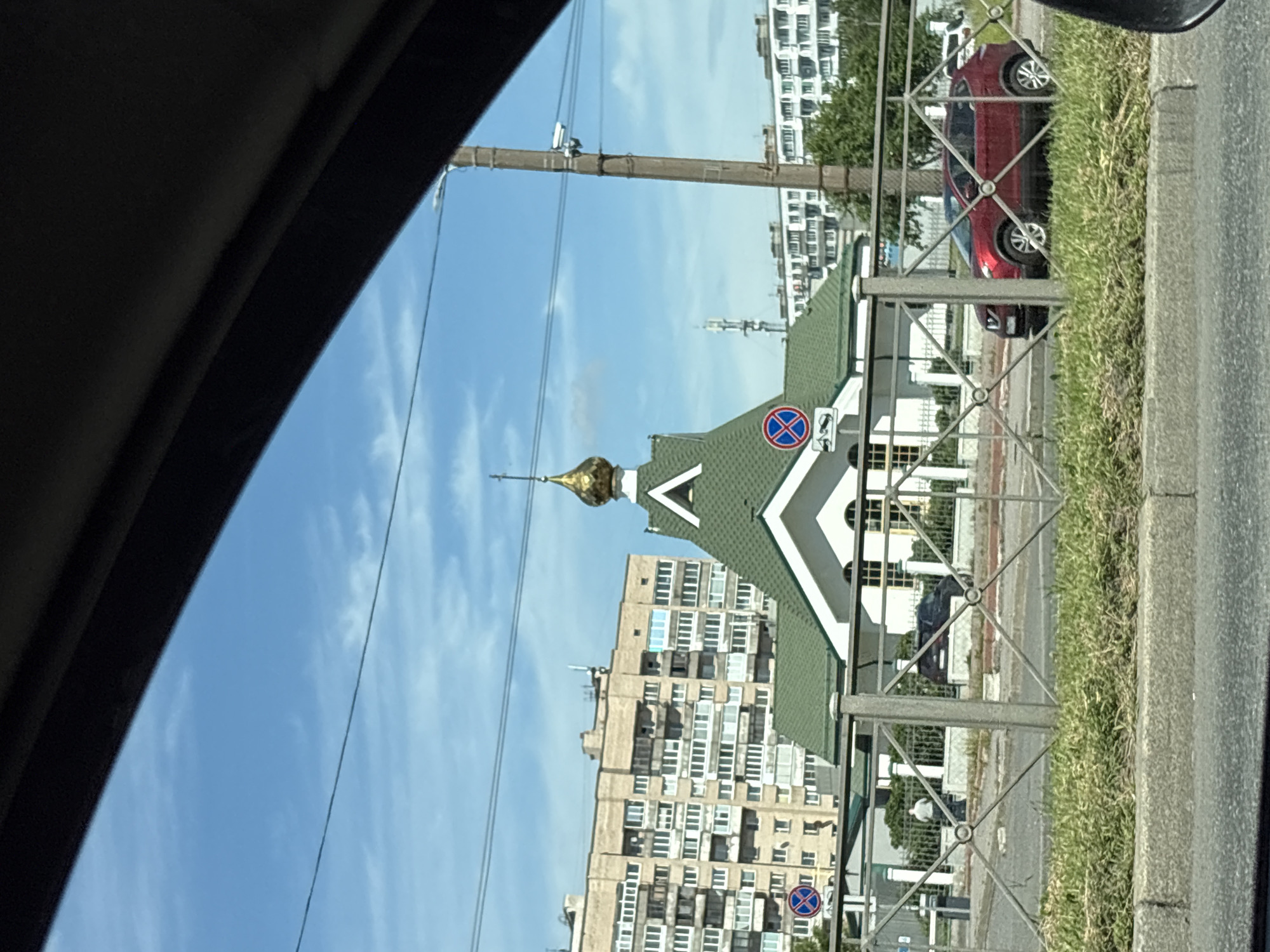 The image depicts a street view in Saint Petersburg, Russia, likely taken from inside a vehicle. The perspective is skewed, suggesting the photo was taken while moving or from a tilted angle. Dominating the scene is a distinctive green-roofed building with a golden dome, characteristic of Eastern Orthodox architecture. Adjacent to it stands a tall, beige apartment building with numerous windows. A red car is partially visible behind a metal fence bordering a grassy area. The sky is blue with scattered white clouds, indicating fair weather during daytime. A utility pole with wires crosses the upper portion of the frame. A traffic sign, a blue circle with a red border and a red diagonal line, is visible near the apartment building. The overall scene suggests a typical urban environment in Russia.