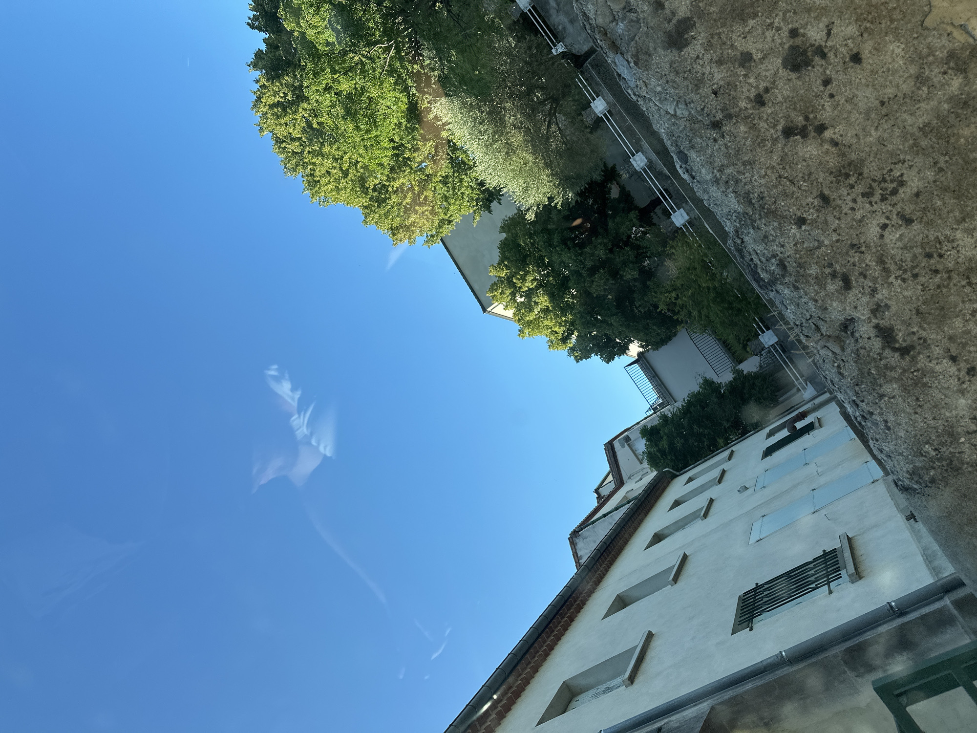 The image, taken from a low angle looking upwards, depicts a section of a building in Anduze, France, under a clear blue sky. The building is a light-colored, multi-story structure with several windows, some featuring light blue shutters. One window has a metal grate. The roofline is visible, adorned with terracotta tiles. Adjacent to the building, lush green trees, including what appears to be an olive tree, add a vibrant natural element to the scene. A stone wall with mossy patches runs diagonally across the foreground, partially obscuring a white railing. The image exhibits a diagonal tilt, giving a dynamic perspective. Reflections are visible in the sky, suggesting the photograph was taken through a window. There are no people or discernible actions depicted.