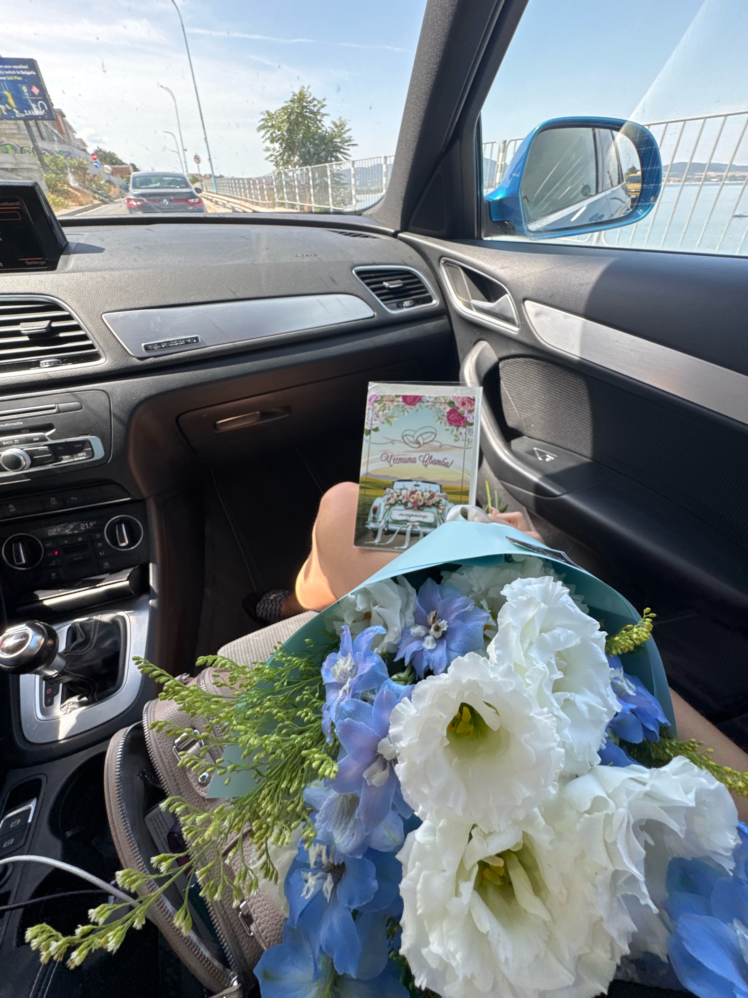 The image shows the interior of a car, specifically the dashboard, passenger seat, and a bouquet of flowers. A wedding card is held in front of the bouquet. The car appears to be in motion, as suggested by the view through the windshield of a road with other vehicles and a body of water to the right. The ambient lighting suggests daytime. The scene takes place in Sozopol, Bulgaria, a coastal city known for its picturesque old town and beaches.