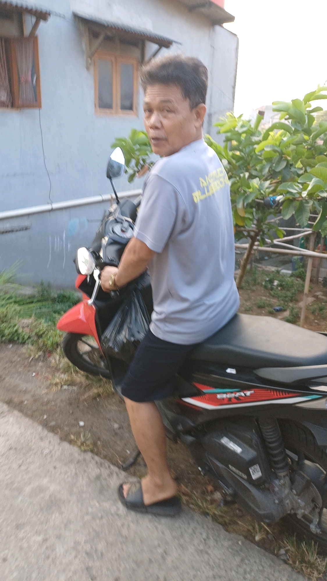 A middle-aged Asian man with dark, styled hair is seated on a red and black motorcycle, looking over his left shoulder. He is wearing a light grey t-shirt with yellow lettering on the back and dark blue shorts. His left hand is on the motorcycle's handlebar, and a gold ring is visible on his left ring finger. A black plastic bag is draped over the front of the motorcycle. The motorcycle has a "BEAT" decal on its side. The man is wearing black slide sandals on both feet. He is positioned on a dirt and gravel path next to a concrete sidewalk. In the background, a pale blue building with two wooden-framed windows and corrugated metal awnings is visible. Green foliage from a tree with broad leaves is to the right of the building and motorcycle. The scene appears to be in Gunung Putri, Indonesia, based on the presence of a "BEAT" motorcycle, commonly sold there.