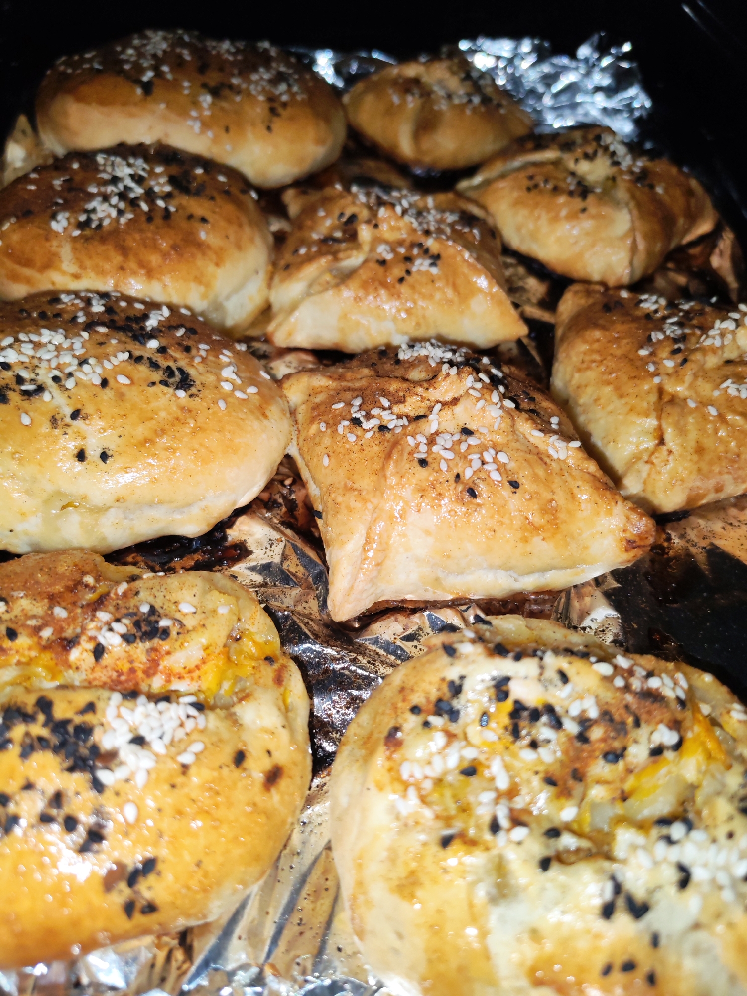 The image shows a close-up of a baking sheet lined with aluminum foil, upon which several baked goods are arranged. There are round pastries and triangular pastries, both glazed with a golden-brown sheen and sprinkled with a mixture of white and black sesame seeds. The pastries appear to be freshly baked, with some areas showing browning and potentially slightly burnt spots. The background is dark and out of focus, suggesting the baking sheet is inside an oven or in a dimly lit environment. There is no indication of Izhevsk, Russia in the image.