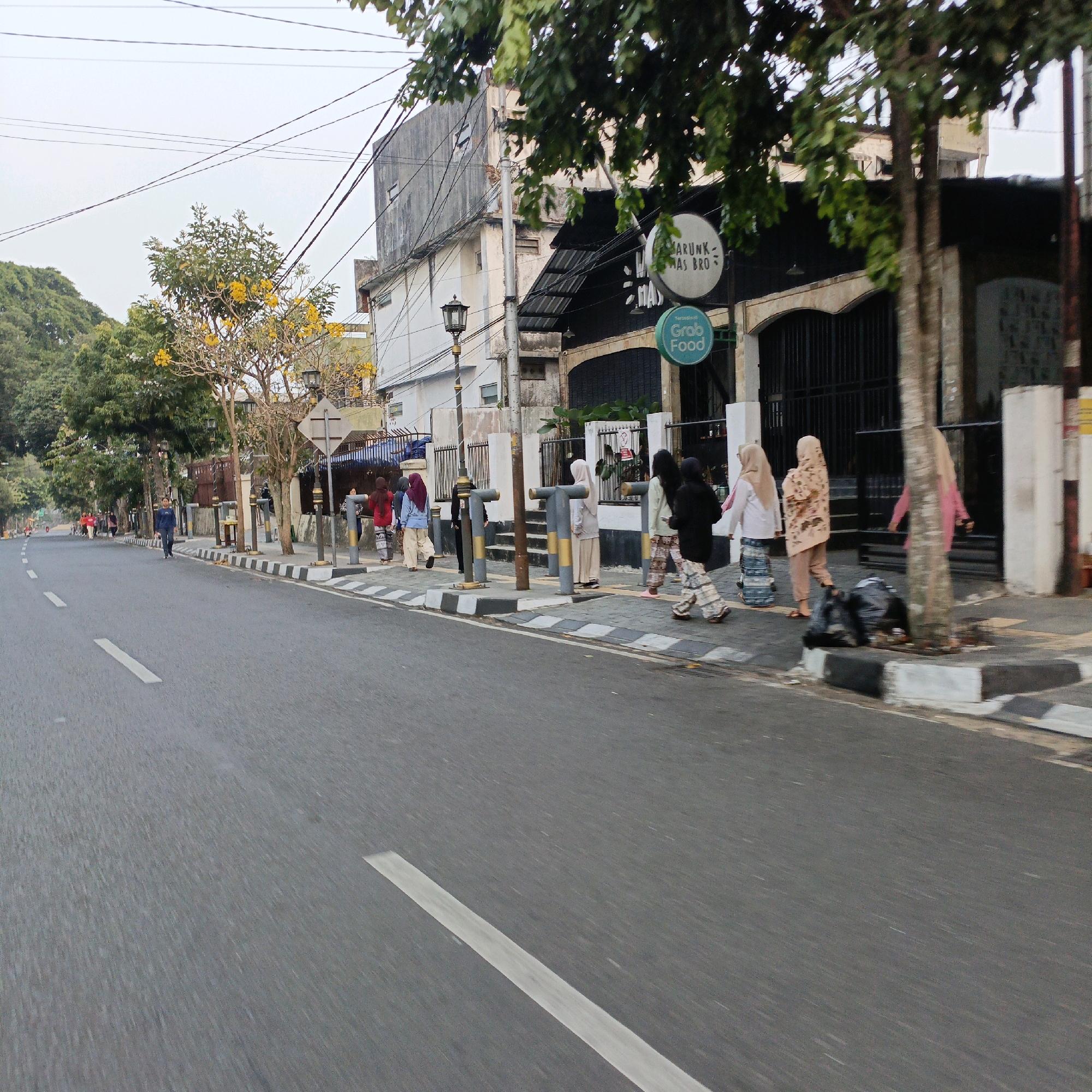 The image depicts a street scene in Salatiga, Indonesia, captured from a moving vehicle. On the right side of the asphalt road, a sidewalk is visible with several individuals walking. Some are wearing hijabs and traditional Indonesian clothing. A tree with yellow flowers is prominent on the left side of the sidewalk. Buildings line the street, some appearing to be commercial establishments, as indicated by signs such as "Grab Food". The sky is overcast. The overall impression is of a typical urban street in Indonesia.