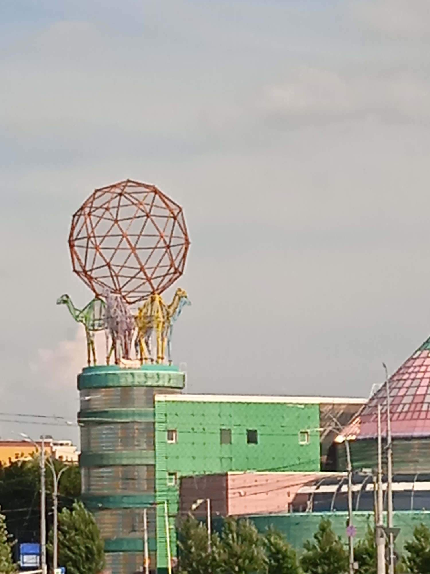 A low-angle, eye-level outdoor daylight medium shot captures a unique architectural sculpture atop a building in an urban setting. The sculpture consists of three stylized, wireframe camels in green, purple, and yellow, standing on a circular platform. Above them, a large, open, geodesic sphere made of thin, interconnected metal rods dominates the sky. The building supporting the sculpture is cylindrical and appears to be clad in green mesh or netting, with horizontal bands of what looks like glass or translucent material. To the right of this, a rectangular green structure with visible windows sits next to a building with a distinctive pinkish-brown lower section and a domed roof with a multicolored, patterned surface. In the background, a clear sky with scattered light clouds is visible. Trees and a utility pole with wires are present in the lower left portion of the frame, and a blue object resembling a truck or container is partially visible on the far left. The overall impression is of a colorful and somewhat whimsical monument on a modern building complex.