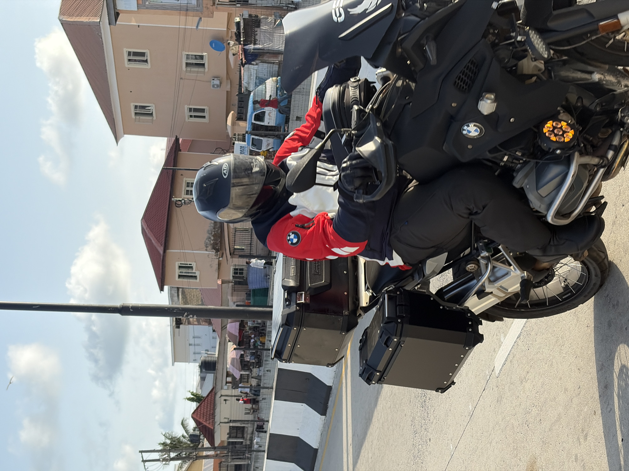 A medium shot shows a person on a black BMW motorcycle on a city street. The rider, wearing a black and red jacket and a black helmet, is looking to the left of the frame. The motorcycle has black storage compartments on the rear. The location is Igboefon, Nigeria; buildings and street life can be seen in the background. A street lamp is positioned to the left of the motorcycle.