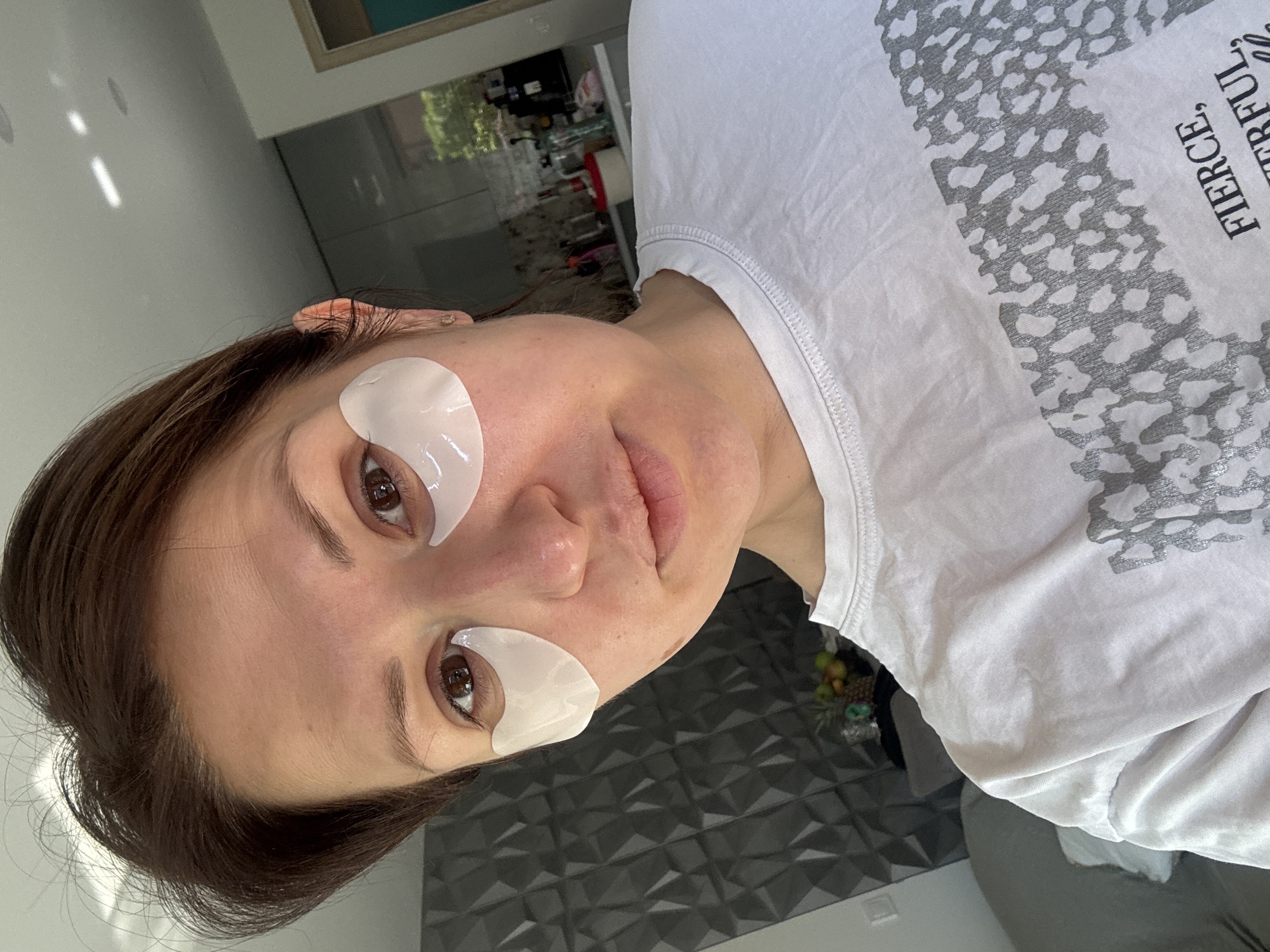 A woman is depicted with eye patches under both eyes. She is wearing a white t-shirt with gray leopard print and the words "FIERCE", "POWERFUL", and "GRATEFUL" printed on it. The background features a blurred interior, including a counter with various items, a textured gray wall, and glimpses of an outside environment. While the location is requested, Sofia, Bulgaria cannot be determined from the visual information alone.
