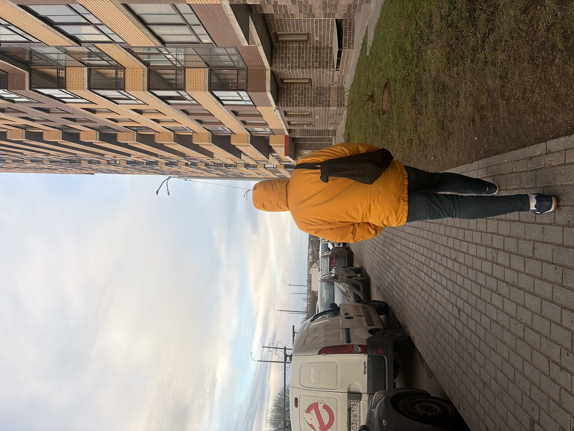 The image depicts a street scene in Saint Petersburg, Russia, with a person wearing an orange jacket walking along a brick-paved sidewalk adjacent to a multi-story building. The building is constructed with light brown and beige brick, featuring a pattern of recessed windows and balcony ledges. A narrow strip of grass and a manhole cover are visible between the building and the sidewalk.
Parked along the street are several cars, including a white van with a Ghostbusters logo and Russian license plate "O03EB 178". The sky is overcast, and overhead power lines are visible. The person is wearing jeans and dark-colored shoes, and a black bag is slung over their shoulder.