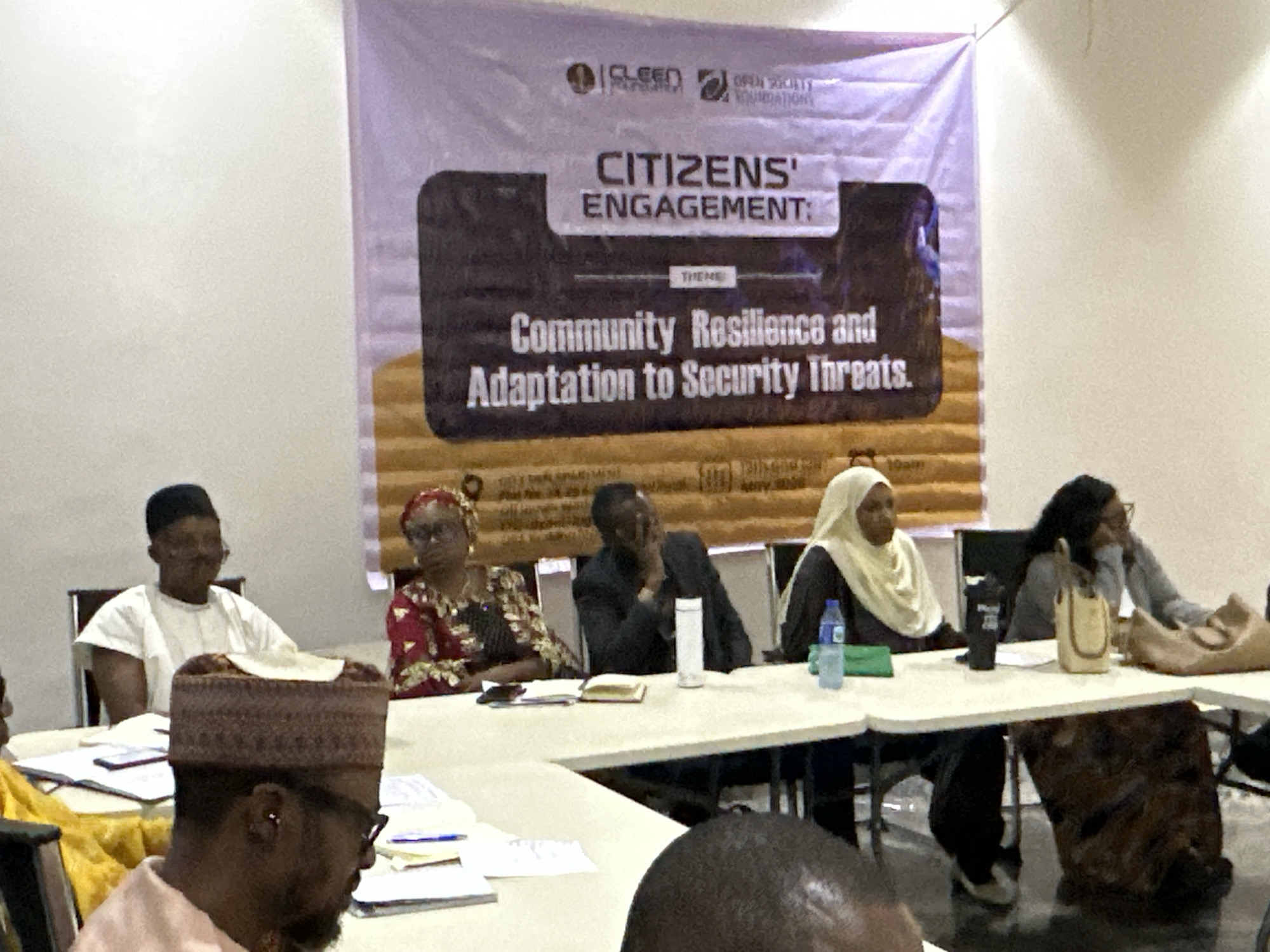 The image shows a group of people seated at a table in front of a banner. The banner reads "CITIZENS' ENGAGEMENT:" followed by the theme "Community Resilience and Adaptation to Security Threats." The banner also includes logos of "CLEEN Foundation" and "Open Society Foundations." The people at the table appear to be attending a meeting or conference. Some are wearing traditional attire, including head coverings. There are papers, notebooks, water bottles, and other items on the table. The location is in the city of Unknown, Unknown.