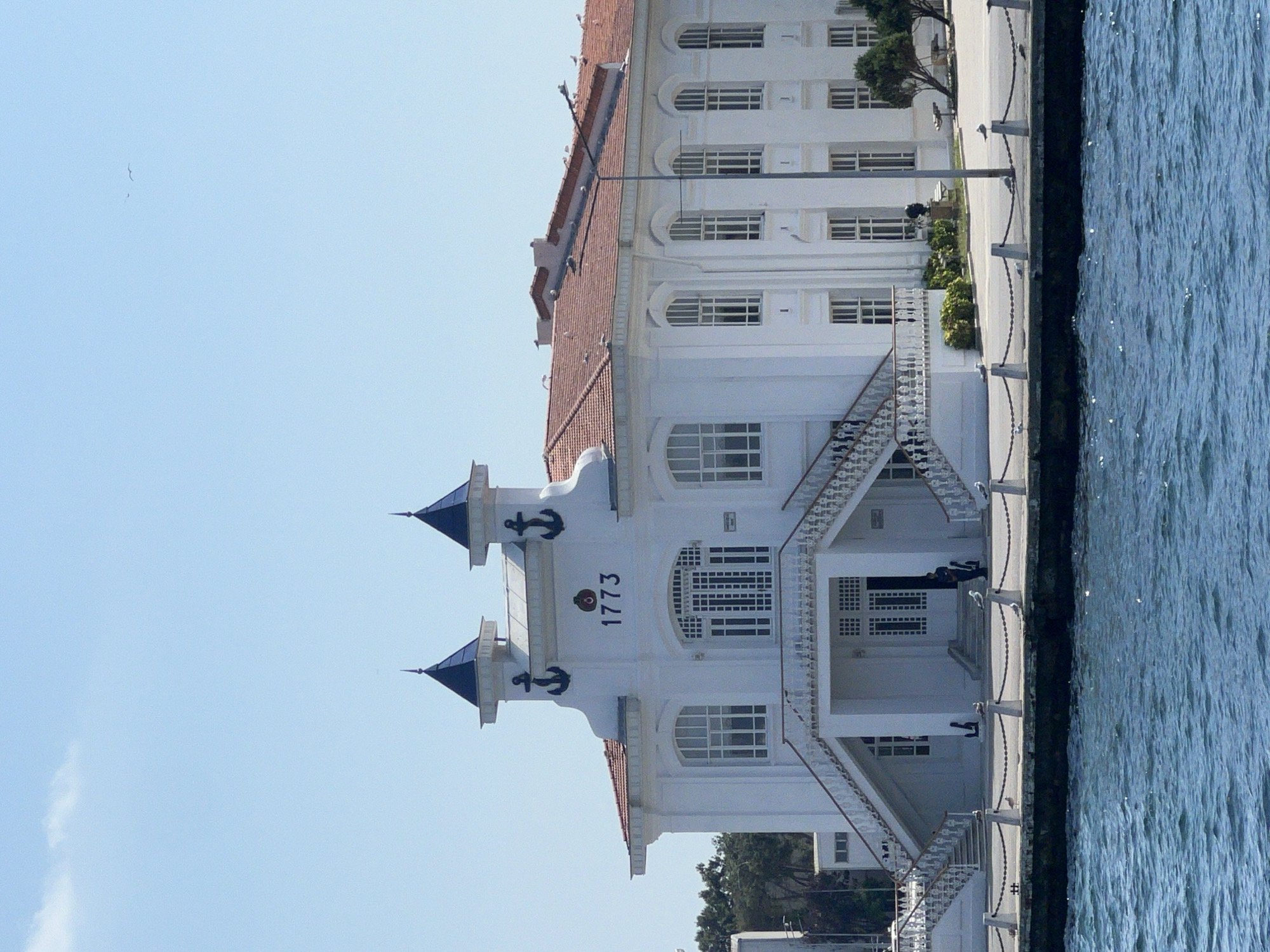 The image shows a white, two-story building with a red tiled roof, situated on the waterfront of the Prince Islands, Turkey. The building has a central section with two prominent turrets topped with dark spires, adorned with anchor motifs. The number "1773" is displayed prominently above the central entrance, along with a circular emblem. A grand white staircase with ornate railings leads up to the main entrance. To the left and right of the central section, the building extends with evenly spaced arched windows on both floors. A chain-link fence with white bollards separates the building from the water. Several seagulls are visible on the roof of the building and along the waterfront. The water in the foreground is choppy and a deep blue. The sky is clear and light blue with a few wispy clouds. There is a street lamp in front of the building.