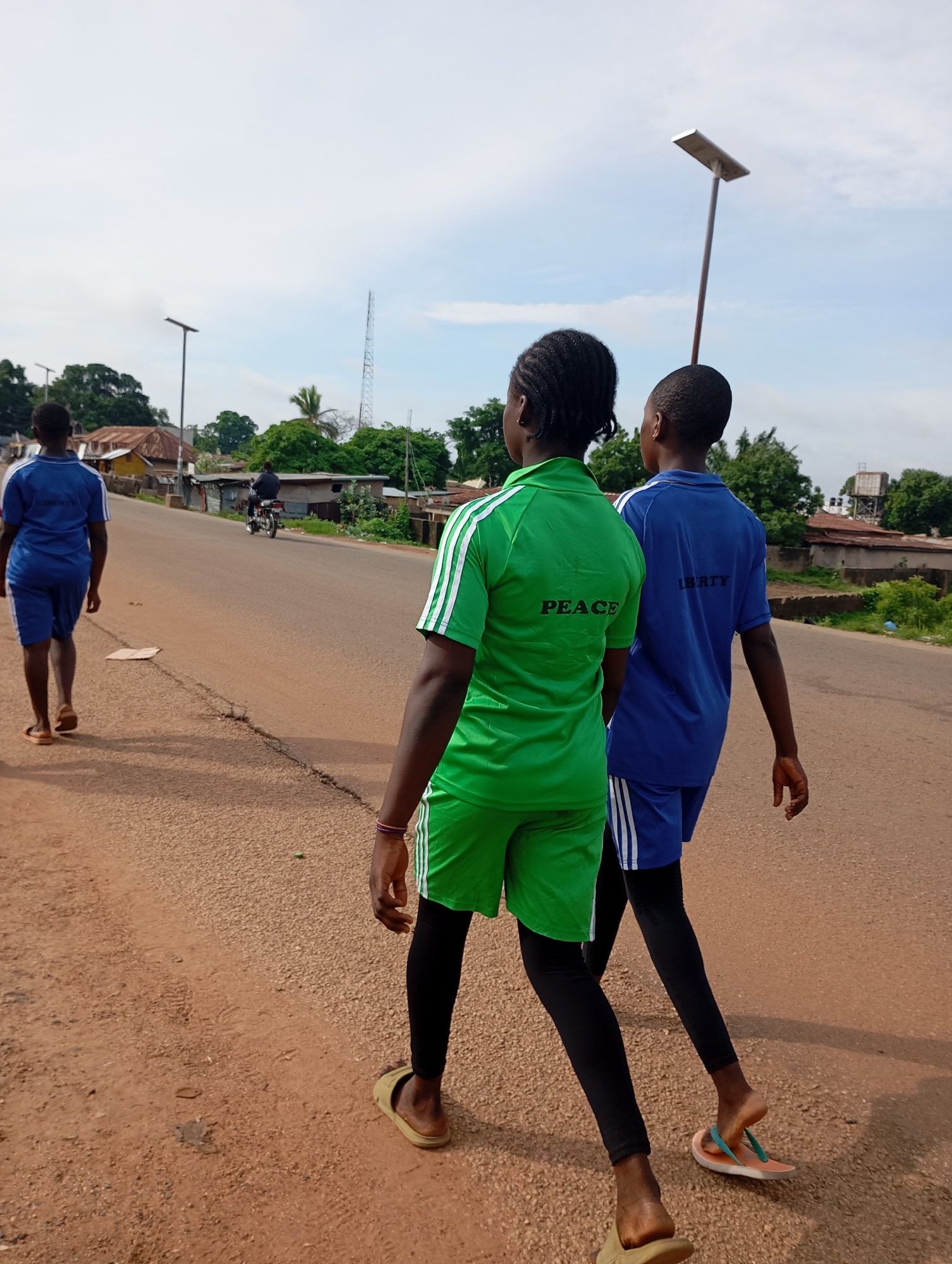 Three individuals are walking on a dirt road in Shendam, Nigeria. The person in the foreground on the left is wearing a blue uniform and walking away from the camera. The person in the middle is wearing a green uniform with "PEACE" printed on the back and walking away from the camera. The person on the right is wearing a blue uniform and walking away from the camera. In the background, there are houses, trees, and a cell tower. The sky is cloudy.