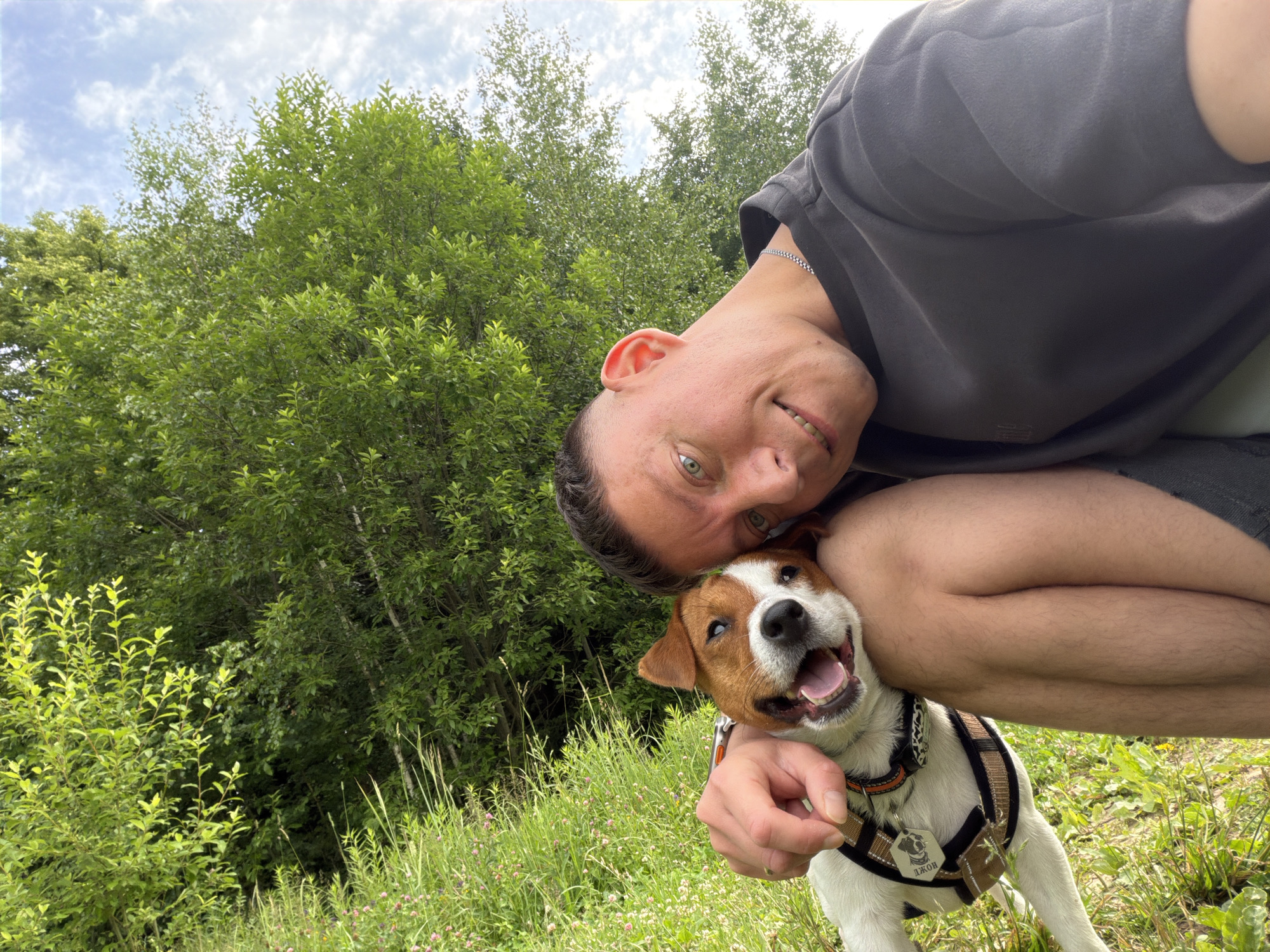 A man and his Jack Russell Terrier are photographed outdoors in a grassy, wooded area. The man is in the foreground, tilting his head down towards the dog and smiling at the camera. He is wearing a dark gray t-shirt and shorts. The dog is held by the man's arm and is looking directly at the camera with its mouth open in a happy expression. The dog is wearing a harness with a tag. The background consists of lush green trees and a cloudy sky. This scene is not identifiable as being in Moscow, Russia.
