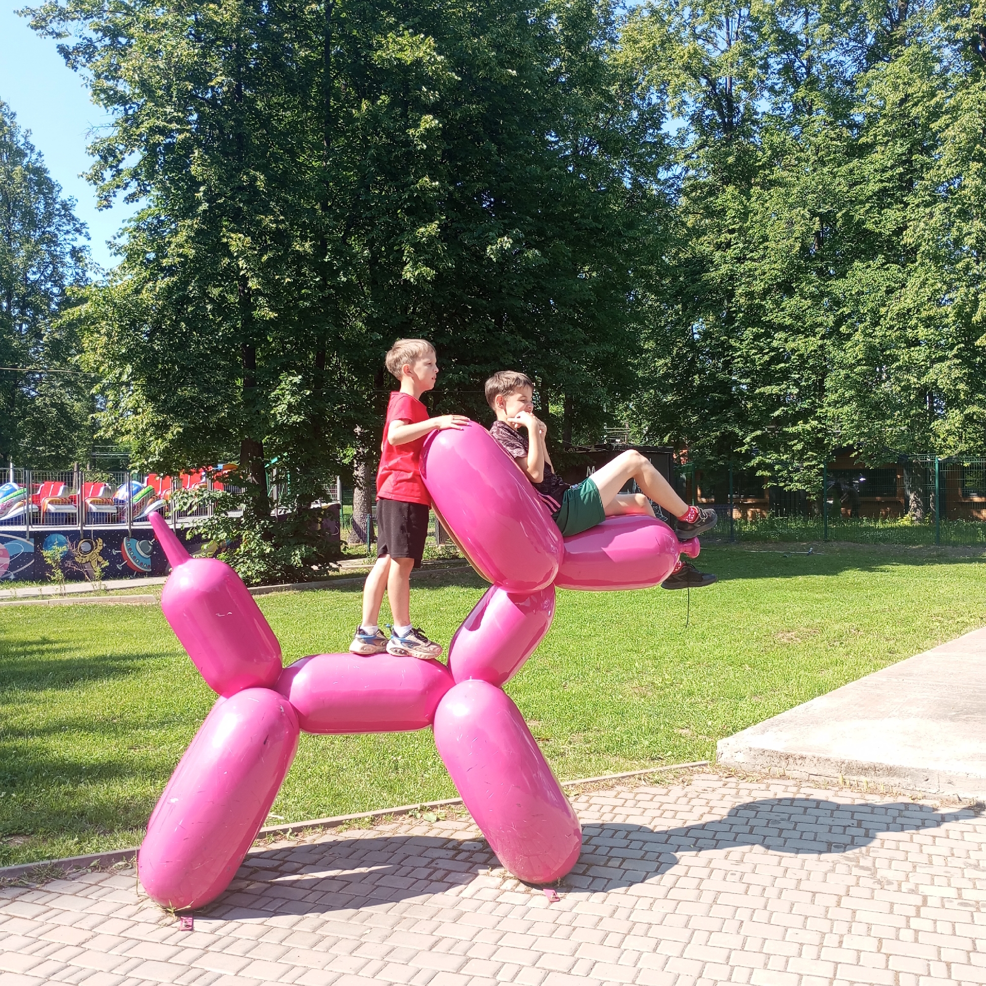 In Izhevsk, Russia, two young boys are interacting with a large, bright pink, balloon-dog sculpture in a park. The sculpture is made of a smooth, glossy material and resembles a stylized dog formed from inflated balloons. One boy, wearing a red t-shirt and black shorts, stands on the sculpture's back. The other boy, in a dark t-shirt and green shorts, is lying on the sculpture's body, propped up on his elbows.
The sculpture is positioned on a paved walkway made of interlocking bricks, with a lush green lawn surrounding it. In the background, large, mature trees with dense foliage create a shaded canopy, suggesting it's a warm, sunny day. Beyond the trees, there's a glimpse of a playground or amusement park area with colorful rides. The scene captures a playful moment in an outdoor recreational space.