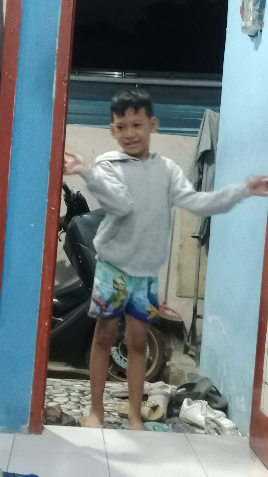 A young boy with short black hair and a light grey hooded sweatshirt and patterned shorts stands with his arms outstretched, facing forward in a doorway. The doorway is framed by a blue wall on the right and a red and blue striped frame on the left. Behind him, a motorcycle is partially visible. Shoes are scattered on the floor in the foreground. The setting appears to be an outdoor covered area, possibly a porch or walkway, with a tiled floor. The background is dark, suggesting it is nighttime or a dimly lit interior. The location is identified as Denpasar, Indonesia.