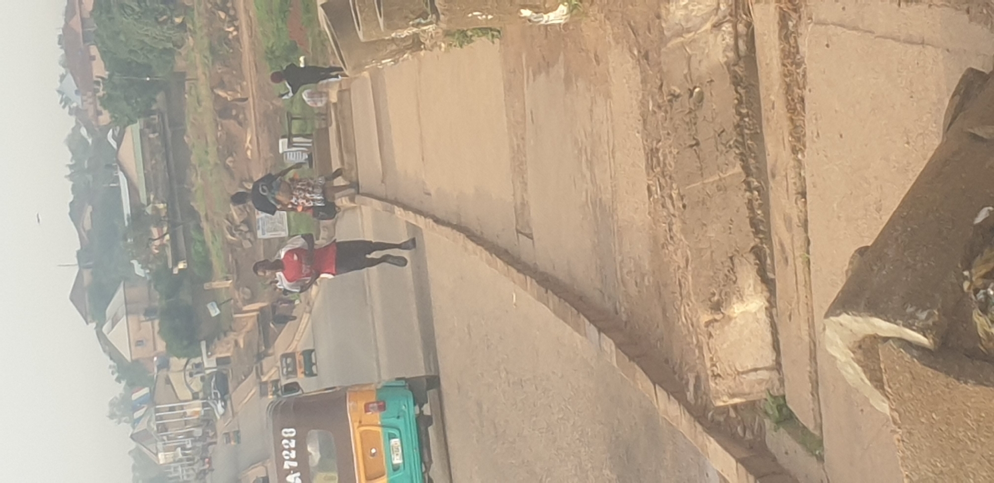 A medium shot captures an outdoor scene on a hazy day, likely in Jos, Nigeria, given the context of the image. In the foreground, a raised concrete walkway runs diagonally from the bottom right towards the center. Beyond this walkway, a paved road curves to the left. On the road, a colorful three-wheeled auto-rickshaw, aqua and brown, with "A 7228" visible on its rear, is parked. To the right of the auto-rickshaw, a young man in a red and white shirt and black shorts is running. Behind him, a woman in a patterned dress is walking, carrying a bundle on her head. Further in the background, more people are visible on the walkway, and a hilly landscape with buildings and vegetation forms the backdrop. The overall atmosphere appears to be that of a bustling urban street.