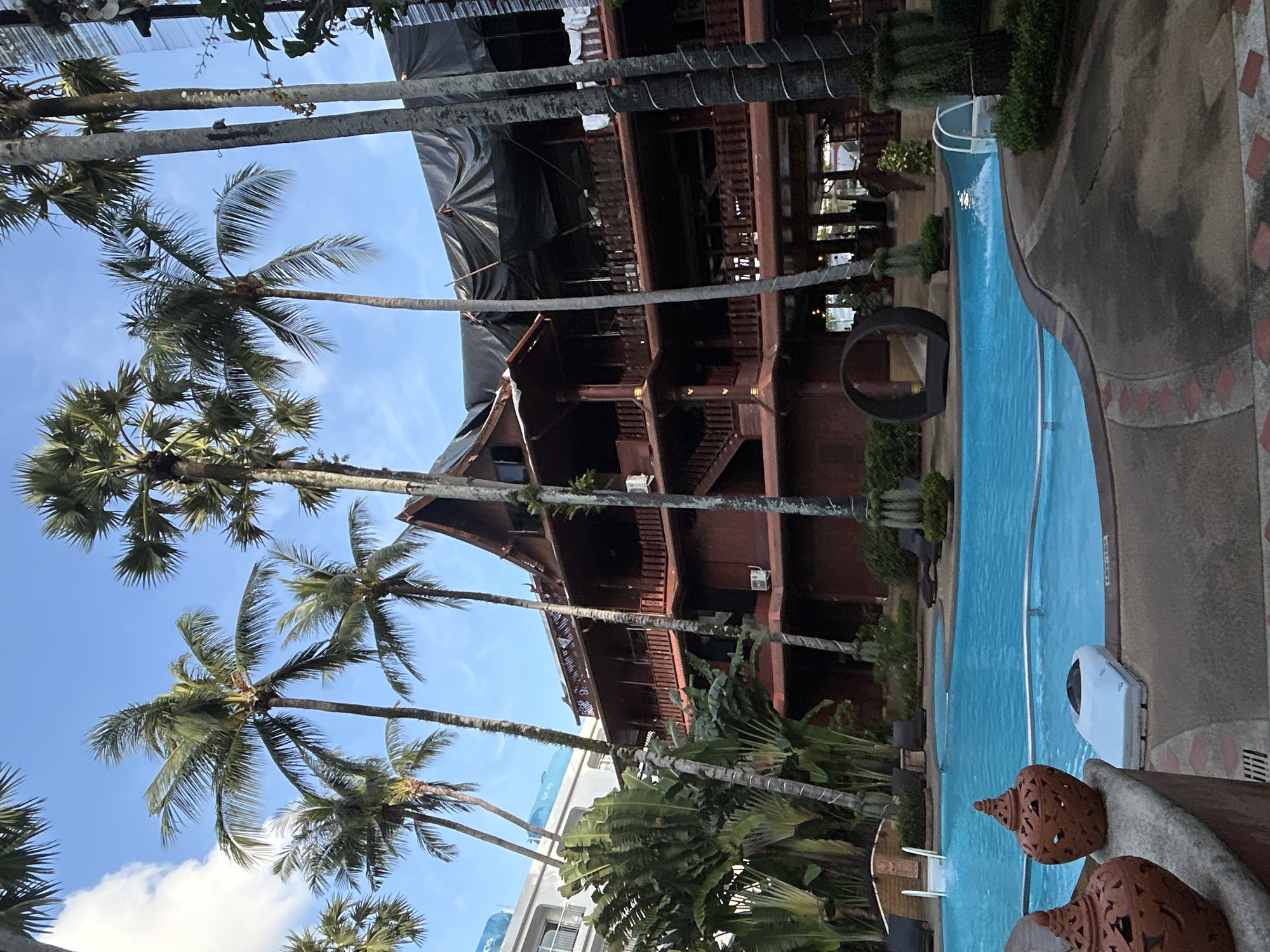 In Patong, Thailand, an outdoor swimming pool with clear blue water is framed by a tropical environment and a multi-story wooden building. Tall palm trees with lush fronds sway against a bright blue sky, with a few white clouds visible. To the left, a modern white hotel building stands partially visible. The wooden building, adorned with balconies and intricate carvings, appears to be under renovation, with a large black tarp covering a section of its roof. Lush green foliage, including banana-like plants, surrounds the pool area. In the foreground, decorative terracotta urns sit on a stone ledge overlooking the pool. A white, oval-shaped object, possibly a spa or jacuzzi cover, is partially submerged in the water. In the middle ground, a unique, circular lounge structure sits near the pool's edge. A person can be seen walking in the distance inside the wooden building.