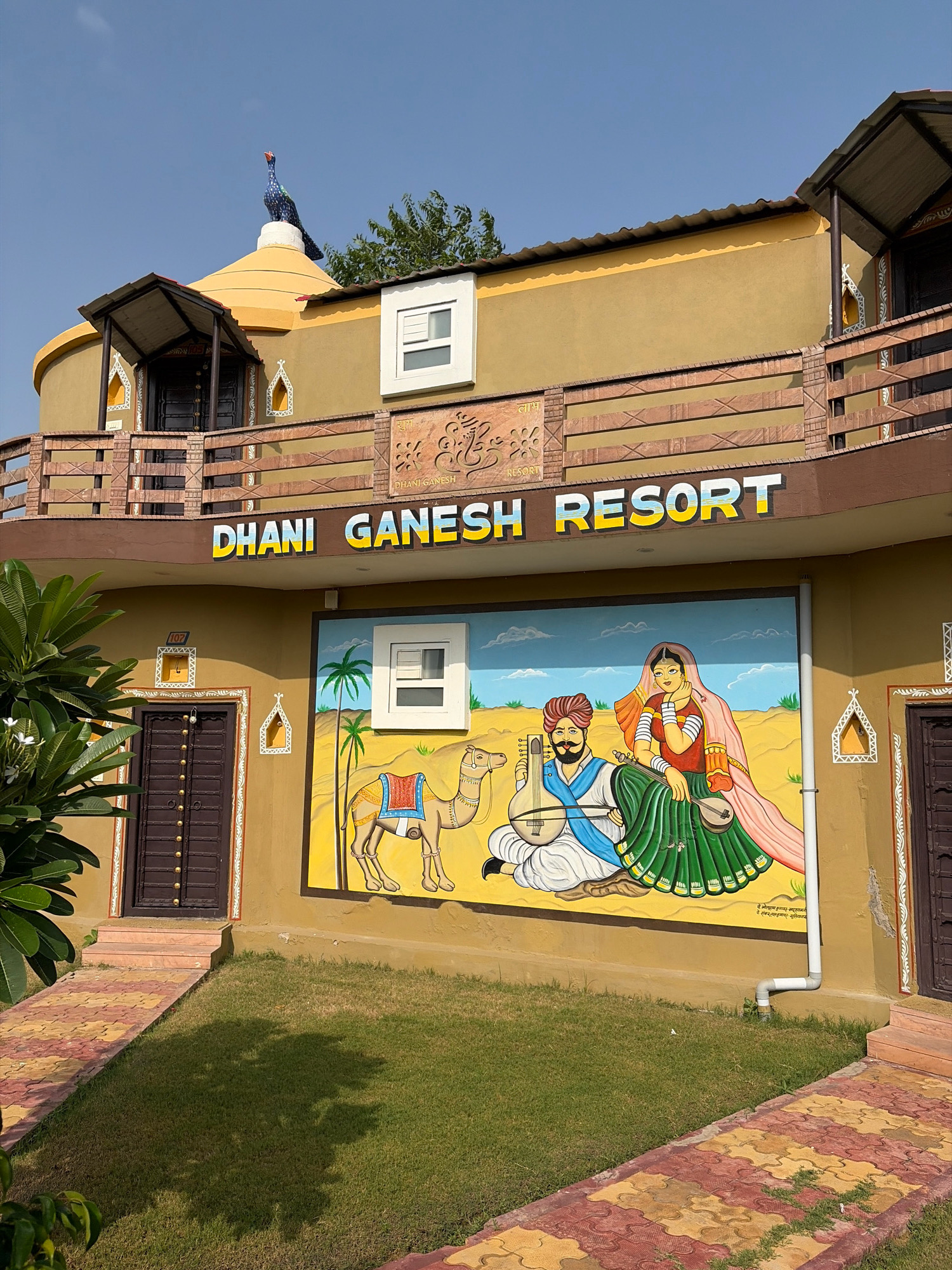 The image depicts the exterior of the "Dhani Ganesh Resort" in Dhokiya, India. The resort features a distinctive architectural style with a mustard-yellow facade adorned with colorful murals. One prominent mural portrays a traditional Rajasthani couple, a man playing a stringed instrument and a woman holding a similar instrument, with a camel standing beside them against a desert backdrop. The resort's name is prominently displayed in large, white and yellow letters above the mural. Other architectural details include a blue peacock statue perched atop a curved, yellow dome, a balcony with wooden railings, and dark brown doors with ornate gold studs. The surrounding environment is a well-maintained green lawn with patterned pathways. The sky is clear blue.