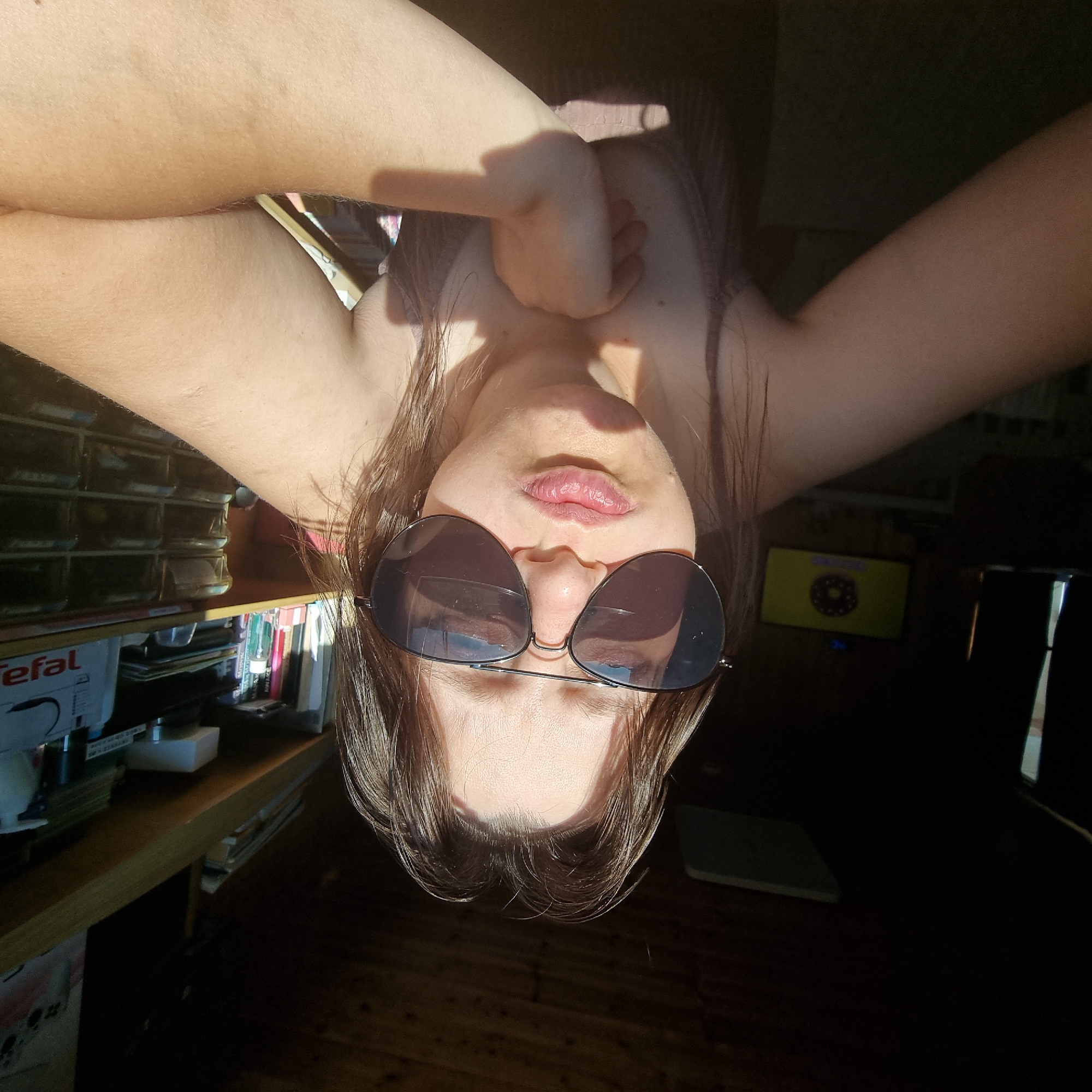 The image shows a young person wearing aviator sunglasses and puckering their lips. They are leaning forward with their arms crossed or resting on a surface. The person has brown hair, and their skin is illuminated by sunlight, casting shadows. In the background, there appears to be a shelf with books and other items, a television screen displaying a circular graphic, and a wooden ceiling. The setting suggests an indoor environment, possibly a room. The image does not provide any information about the city of Dunnae-myeon, South Korea.