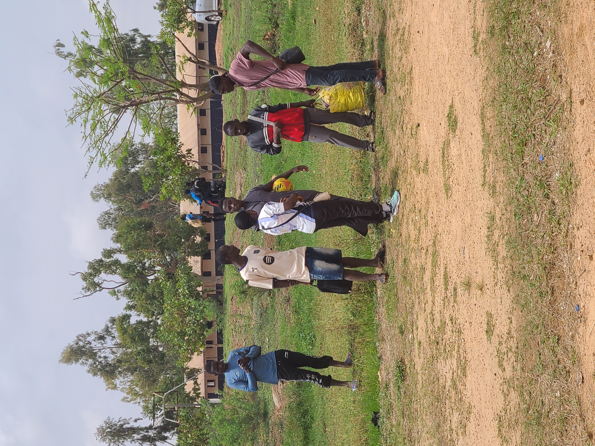 Five young men stand on a grassy field next to a dirt path. Behind them is a row of buildings, likely in Bukuru, Nigeria. The first man wears a blue shirt and dark pants. The second wears a light-colored shirt and denim shorts. The third holds a yellow soccer ball. The fourth wears a red jacket. The fifth carries a yellow bag. The sky is overcast. Trees border the field.