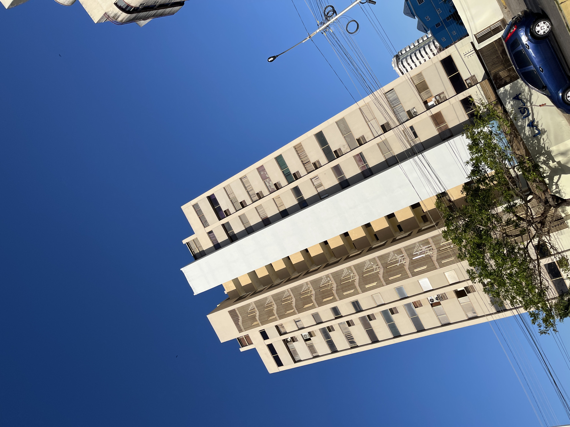 The image features an upward-looking view of an urban landscape under a clear blue sky, with buildings and street elements oriented vertically on the right side of the frame. A large expanse of deep blue, cloudless sky fills the left side and a portion of the top and bottom of the image, with a tiny dark speck, possibly a bird, visible in the upper-left sky area.
Dominating the right side of the frame is a tall, light-colored multi-story building, extending from the top to the bottom of the image. This building is characterized by numerous rectangular windows, some appearing dark, others reflecting light. A prominent, broad white architectural band or balcony spans horizontally across the building's mid-section. Below this band, another section of the building has recessed areas and a decorative facade with horizontal slats and a pattern resembling palm trees.
Further to the right, a dark blue car is parked on what appears to be a street or sidewalk, positioned vertically in the frame. Adjacent to the car, a lush green leafy tree is partially visible, casting distinct shadows on the ground. On a wall behind the car and tree, the white text "LA CASA" is clearly visible against a darker background.
In the upper-right corner of the image, above the main building and tree, a street light pole with a black lamp fixture extends diagonally, accompanied by a tangle of black electrical or utility wires crisscrossing the frame. Other buildings are partially visible: a light-colored structure with curved architectural details in the very top-right corner, and a blue building with a white horizontally-striped building beneath it, both seen above the main building on the right side of the frame.
The scene is brightly lit by direct sunlight, casting sharp shadows, indicating a clear daytime, likely midday or early afternoon. The setting is an urban environment, consistent with Porlamar, Venezuela. No specific activity or people are visible.