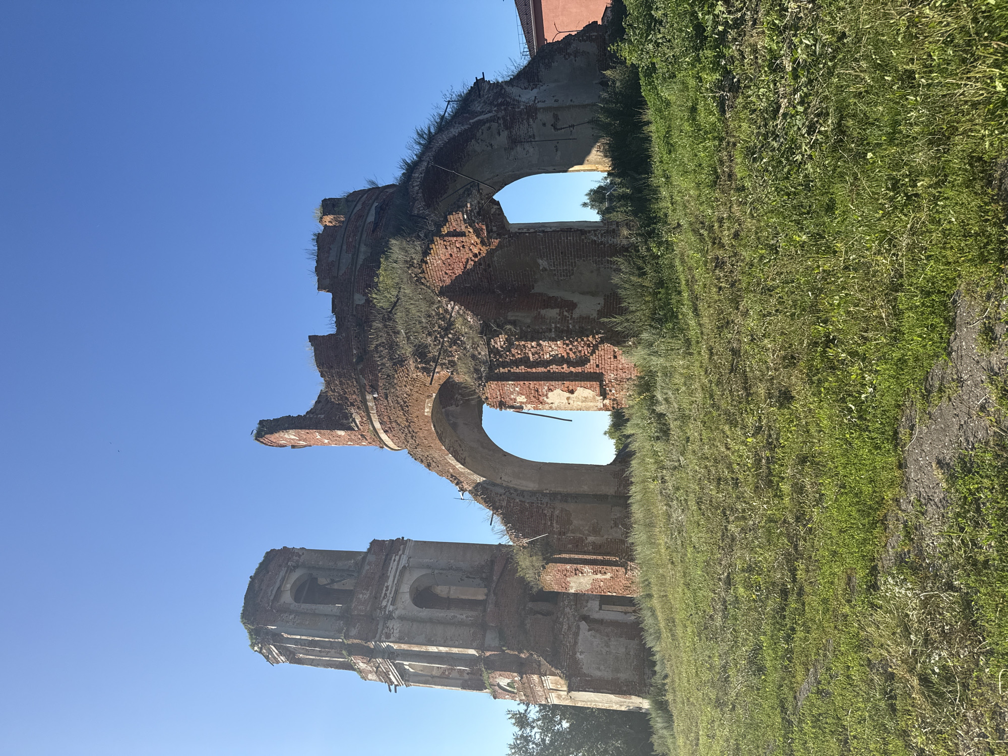 An outdoor, eye-level, medium shot captures the ruins of a brick structure against a clear blue sky. The left side of the frame is dominated by a tall, weathered brick tower with arched openings. To its right, the crumbling remains of a circular or semi-circular structure exhibit large arched voids. Overgrown grass and weeds cover the ground and creep up the sides of the ruins. In the far right background, a portion of a modern building with a red roof is visible. The scene is bathed in sunlight, casting subtle shadows.