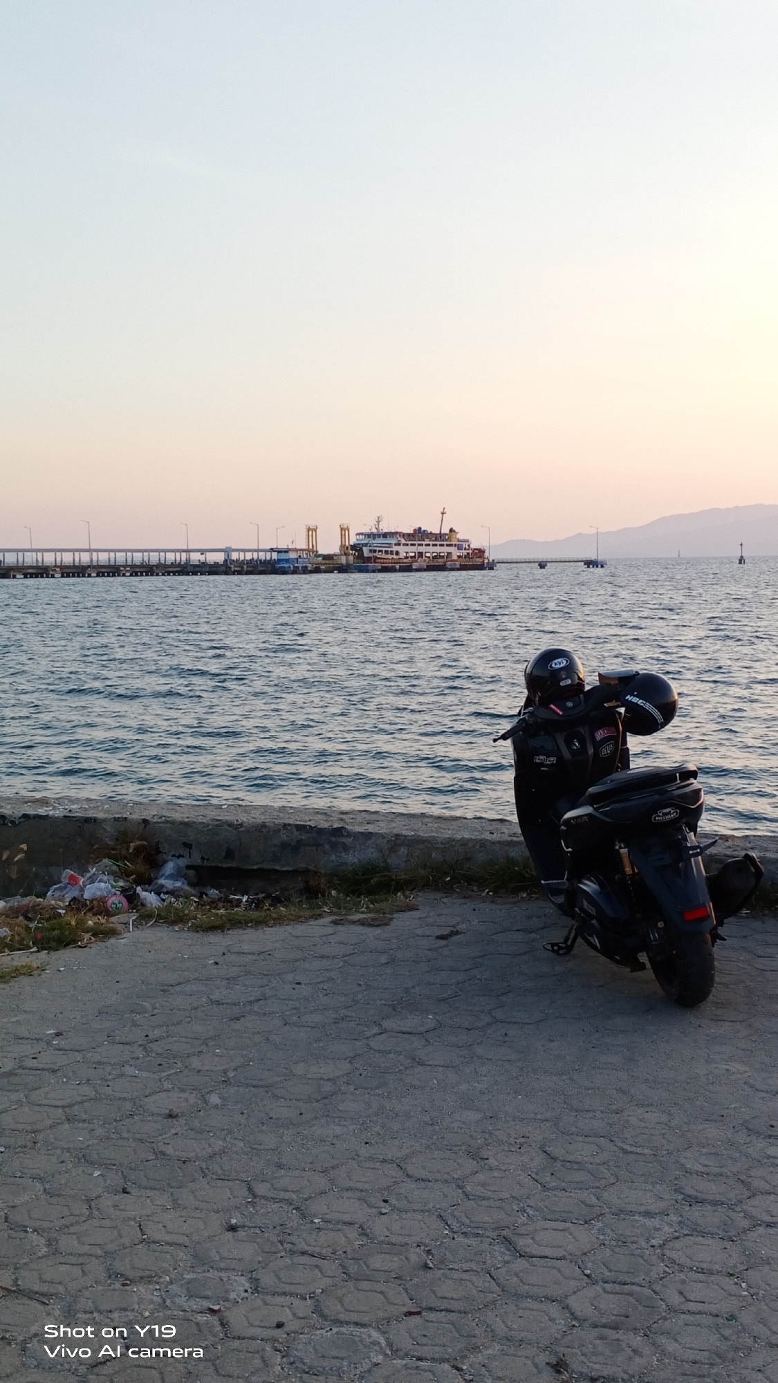 An evening view captures a serene seascape with a ferry docked at a distant pier in Sea, Indonesia. The sky transitions from a pale blue to a soft orange, hinting at sunset. The calm sea reflects the pastel hues, with gentle waves lapping at a concrete barrier in the foreground. A black motorcycle, equipped with two helmets, is parked on the textured pavement. To the left of the motorcycle, scattered trash lies near the concrete barrier, contrasting with the natural beauty of the scene. The pier extends horizontally across the frame, supporting a long line of streetlights and a ferry, its lights reflecting on the water. The background reveals distant hills bathed in the warm light of the setting sun.