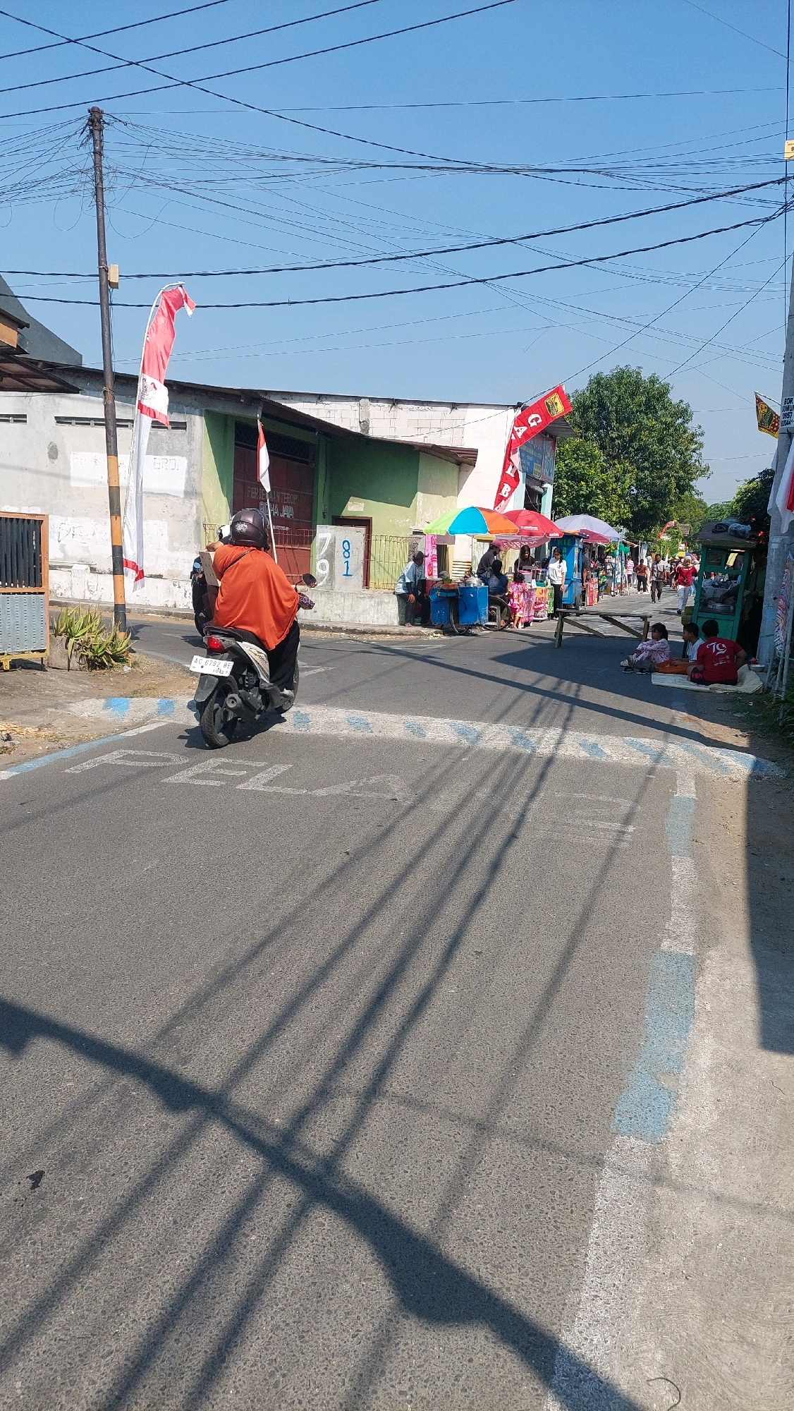 The image depicts a street scene in Sidoarjo, Indonesia, on a clear, sunny day. The road is paved, with blue and white markings indicating a pedestrian crossing. In the foreground, a person is riding a motorcycle away from the camera, wearing an orange cloak and a helmet. The motorcycle has a license plate that reads "AC 0792 BE".
To the left of the road, there is a building with a gray facade and a green-tiled roof, and a white pole with red and white Indonesian flags flying. There are also some potted plants.
On the right side of the road, there are several street vendors with colorful umbrellas selling goods. A few people are gathered around the stalls, and some children are sitting on the ground, possibly waiting or resting. In the background, more people can be seen walking along the street, and trees and other buildings are visible. Overhead, a complex network of power lines stretches across the sky. The overall atmosphere is that of a bustling local street on a bright day.
