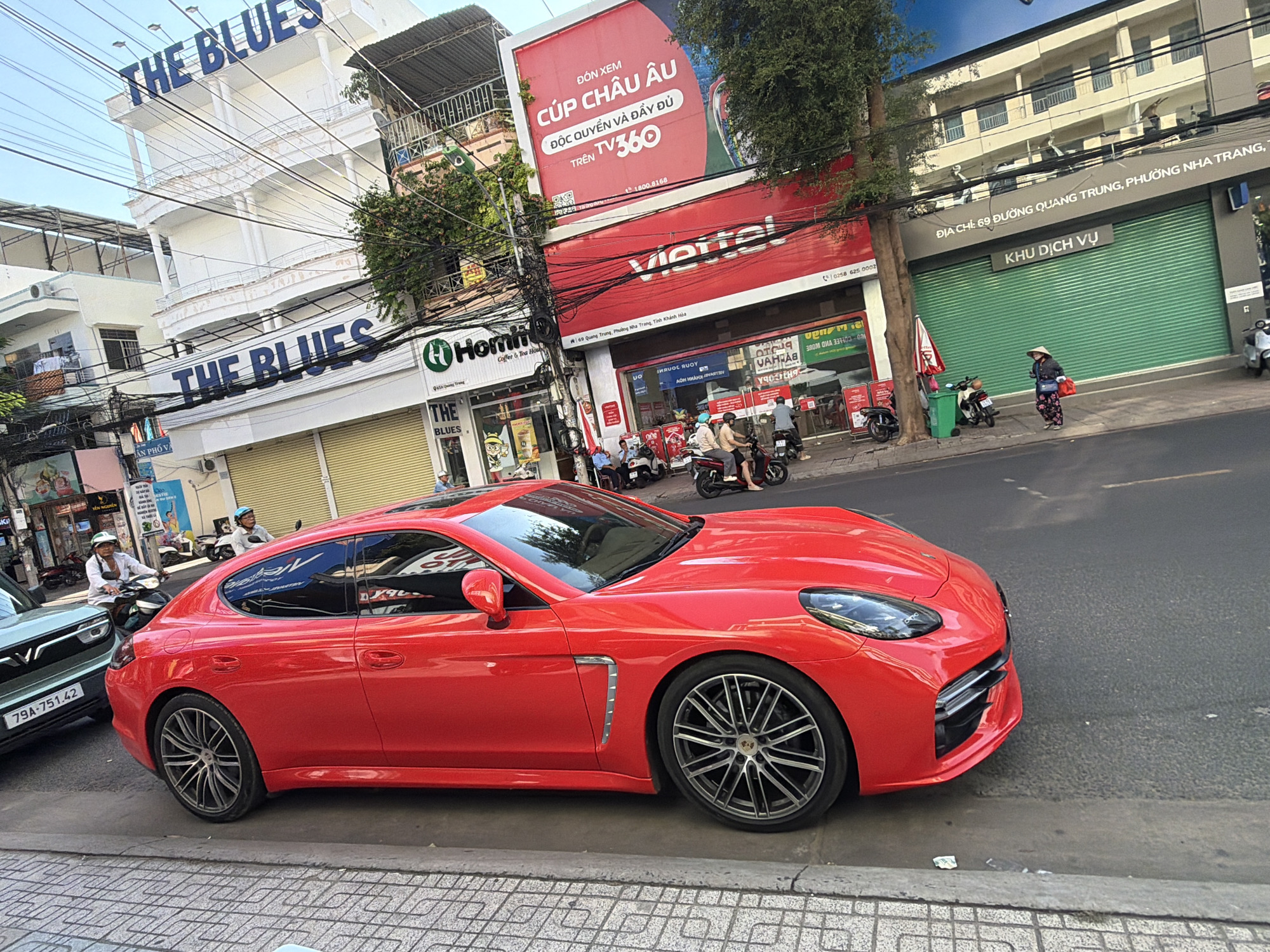 The image captures a street scene in Nha Trang, Vietnam, featuring a vibrant red Porsche Panamera parked on the right side of a paved road, adjacent to a sidewalk. The car is positioned in the foreground, taking up a significant portion of the frame. The sidewalk is constructed with a patterned tile design.
Behind the car, a line of buildings is visible. To the left, a white building with prominent blue signage reads "THE BLUES" on multiple levels. Further to the right, a red building displays a large advertisement for "Viettel" with Vietnamese text and a television screen graphic. Another section of the building shows "CÚP CHÂU ÂU" which translates to "European Cup".
Several people are present in the scene. A person wearing a conical hat is walking on the sidewalk to the right of the red Porsche, carrying a blue bag and a red bag. In the background, there are individuals on motorcycles and scooters, suggesting ongoing street activity. A light green car with a license plate "79A - 751.42" is parked to the left of the red Porsche.
The environment appears to be a typical urban street with overhead electrical wires crisscrossing the sky. Trees are also present, with foliage visible on the left and right sides of the frame. The overall impression is a snapshot of everyday life in a Vietnamese city, with modern vehicles and commercial establishments.