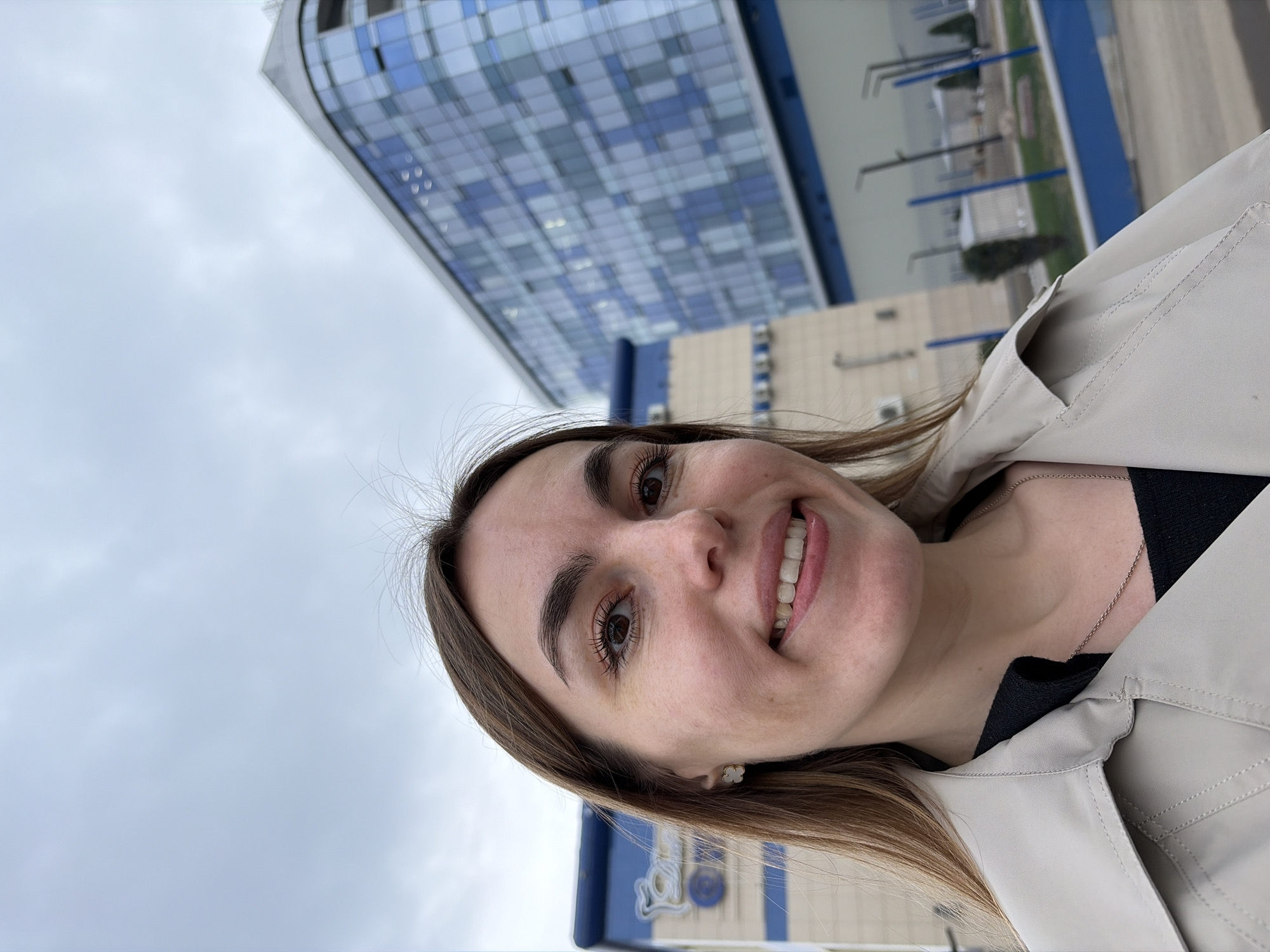 A woman is smiling at the camera. She is wearing a light-colored jacket over a black shirt, with a silver necklace visible. The background features a modern building with blue and white glass panels, characteristic of contemporary architecture. The sky is overcast. The location is in Izhevsk, Russia, as identified by the architectural style of the building in the background.