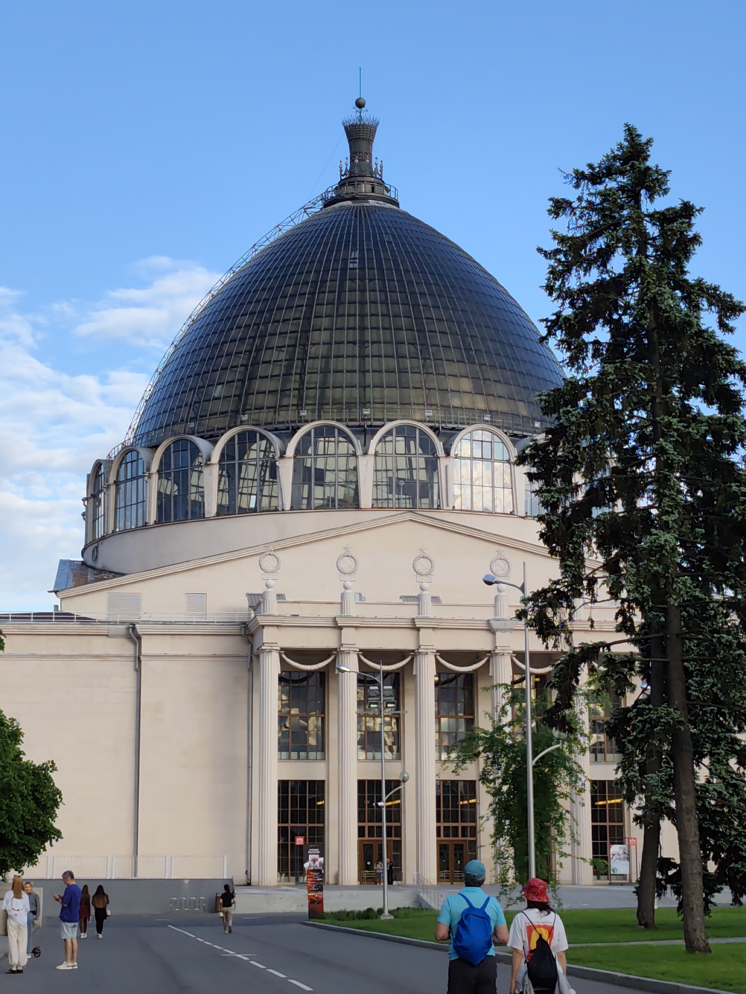 The image depicts a building in Moscow, Russia, likely the Cosmos Pavilion at the VDNKh (Exhibition of Achievements of National Economy). The building features a large, glass-paneled dome on top of a neoclassical structure with columns and arched windows. Several people are walking on the street in front of the building, and trees are present in the foreground on the right. The sky is partly cloudy.