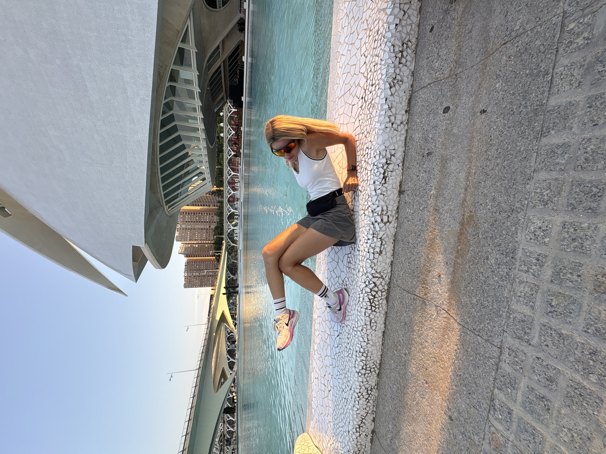 In Valencia, Spain, a blonde woman with sunglasses is lying on a mosaic-covered ledge by a turquoise pool. She is wearing a white tank top, grey shorts, and athletic shoes. The background features the futuristic architecture of the City of Arts and Sciences, including the Hemisfèric building and a modern bridge. The sky is clear and blue, and the sun is casting long shadows.