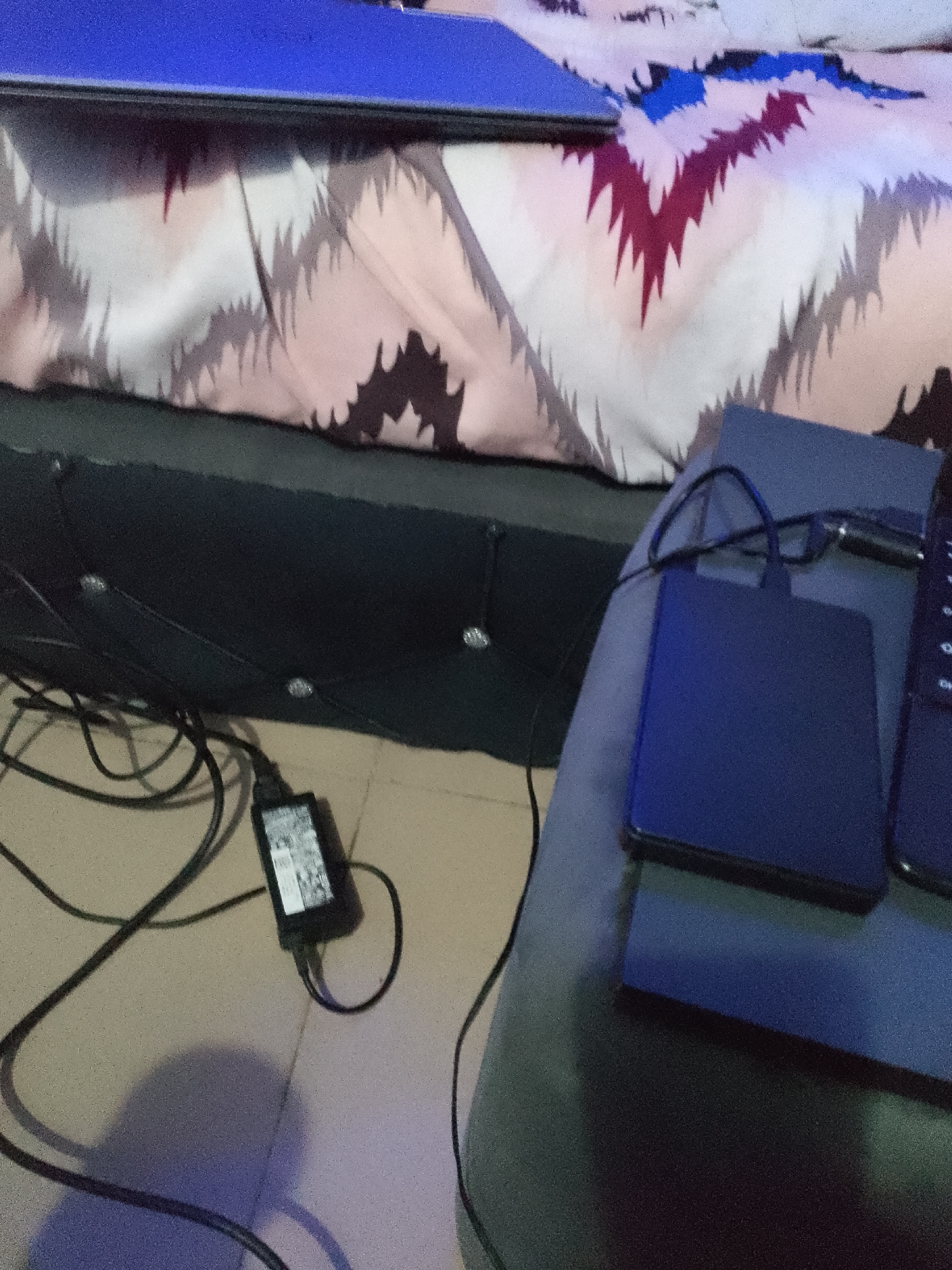 A close-up, high-angle shot shows a bed with a patterned comforter in the background. In the foreground, a black power adapter with a white label is connected to a black cord, which is draped across the floor. A dark blue external hard drive is positioned on a dark surface, and part of a laptop and a smartphone are also visible on the same surface. The scene appears to be in a bedroom.