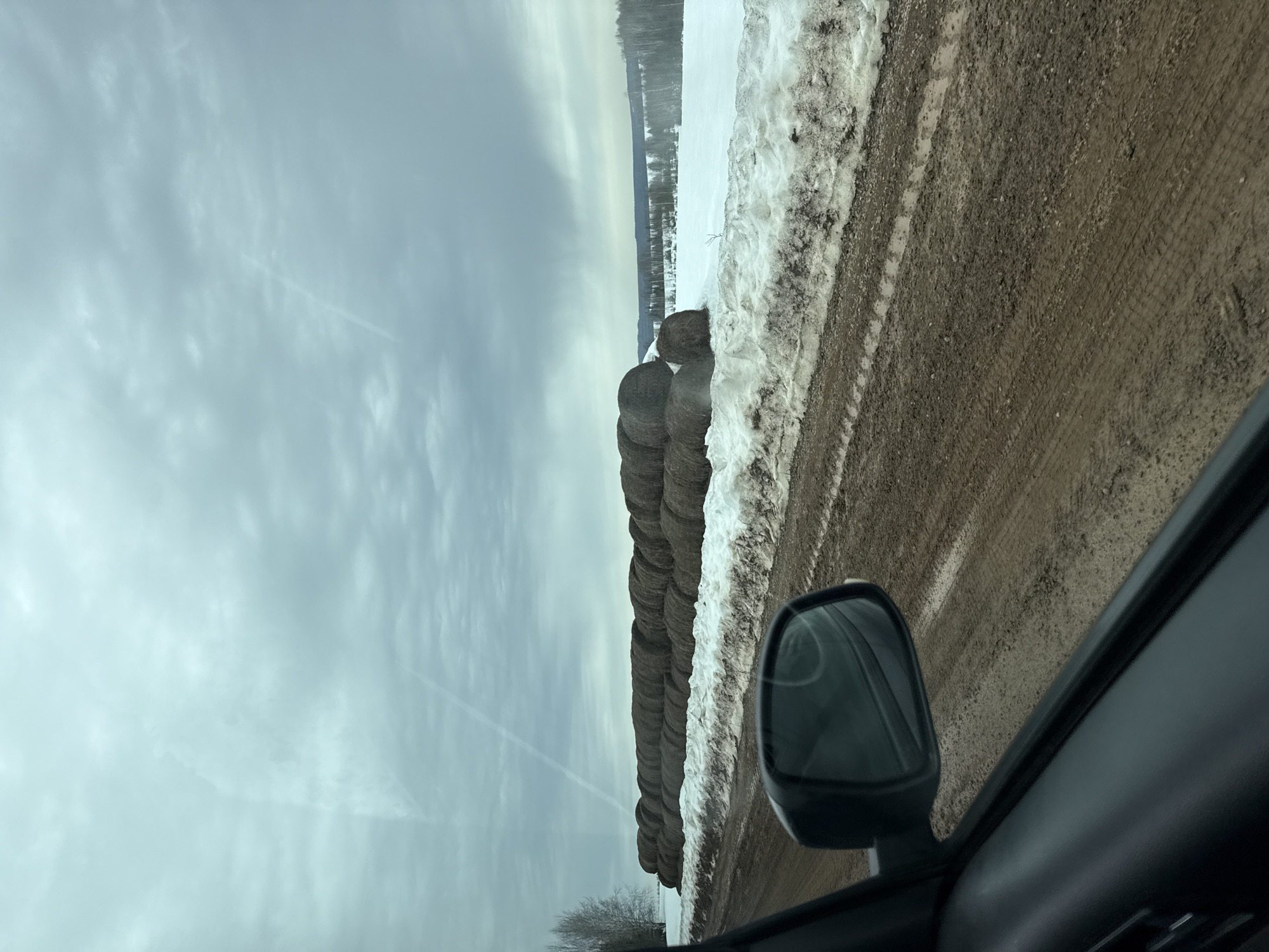 The image shows a scene from a car's perspective, likely on a rural road. A row of hay bales runs alongside the road, partially obscured by snow. Beyond the bales, a snow-covered field stretches towards a distant treeline. The road surface is muddy and shows tire tracks. The sky is overcast with light, streaky clouds. The car's side mirror is visible in the foreground, suggesting the photo was taken while driving. The location is reported to be in Малая Уса, Russia. There are no visible people in the image.