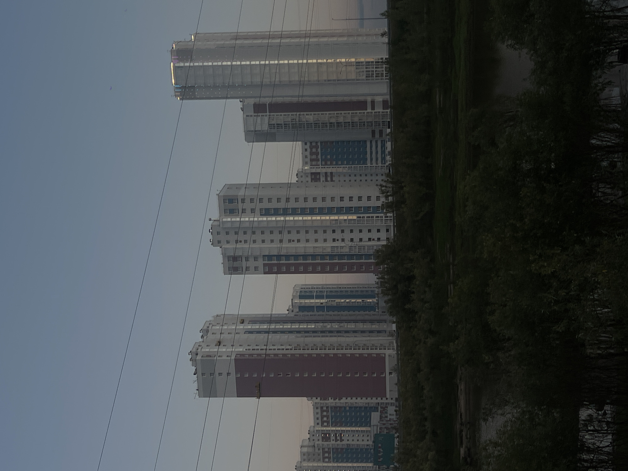 The image shows a line of modern, multi-story residential buildings in Surgut, Russia, under a twilight sky. The buildings are primarily white and light-colored, with some featuring maroon and blue paneling. Several power lines are visible in the foreground, stretching horizontally across the frame. Below the buildings, a dense band of dark green foliage, likely trees and bushes, obscures the lower part of the structures and extends towards a dark body of water, possibly a river or lake, which is partially visible in the bottom left of the image. The overall scene is one of urban development integrated with natural elements, captured during the transition from day to night. No people are discernible in the image.