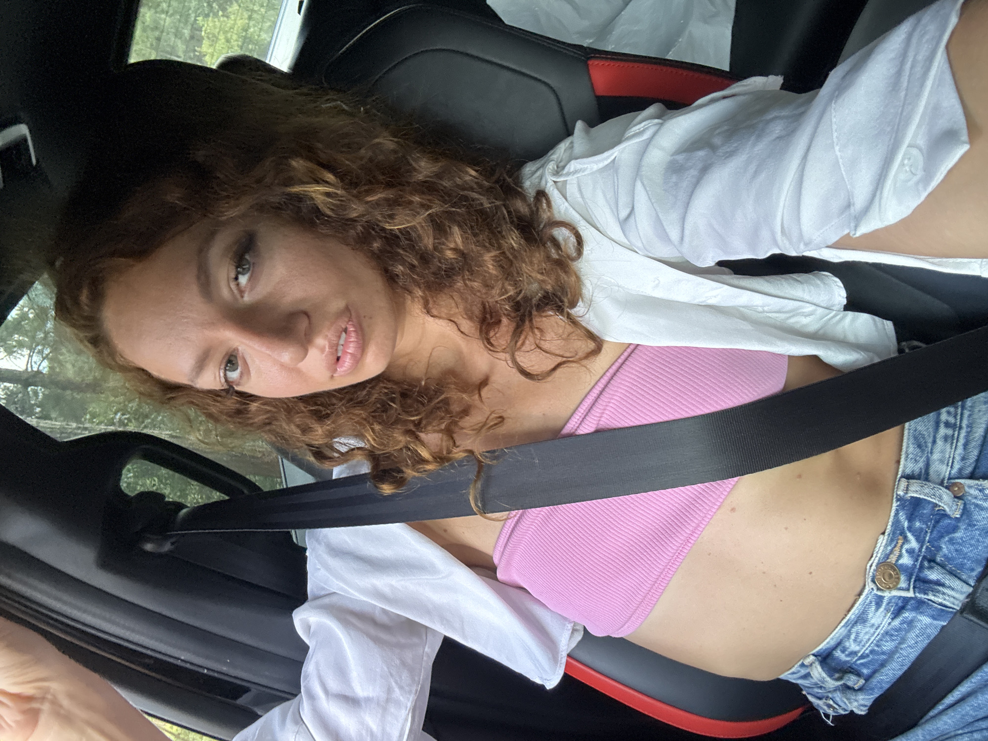 A woman with curly brown hair and fair skin is in a car, wearing a pink top, a white open shirt, and blue jeans. She is wearing a seatbelt. The car's interior is black with red accents. Outside the car window, trees can be seen. The location is not possible to determine as Ngã Ba Kim, Vietnam.