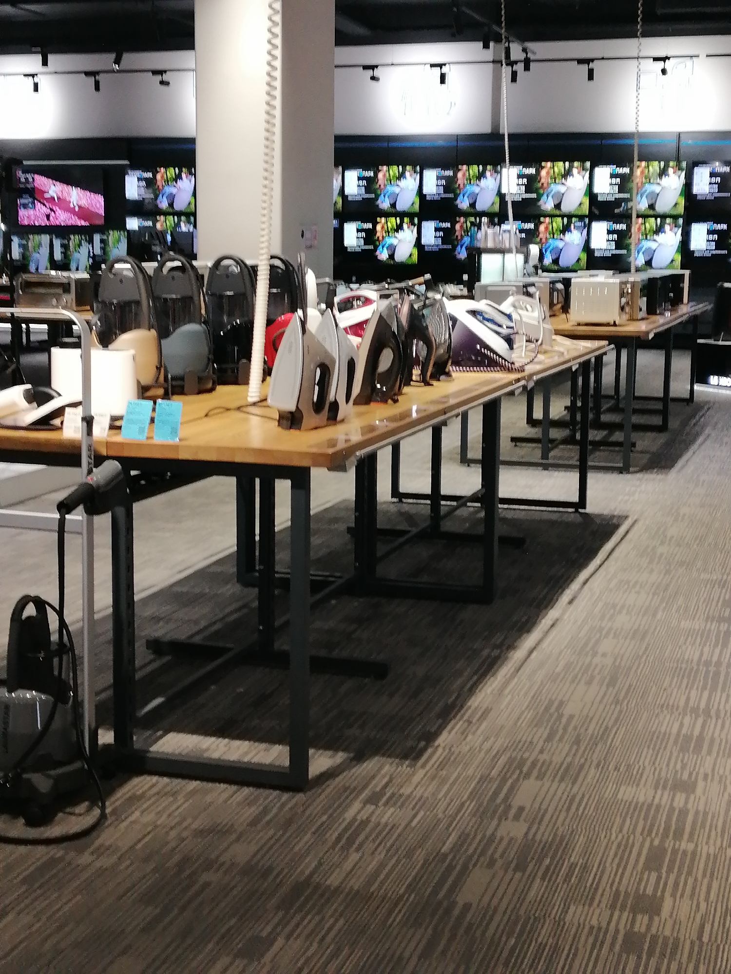 The image shows a retail store interior with a focus on home appliances, specifically irons. In the foreground, several irons are displayed on a wooden table with dark metal legs. The irons vary in color and design, with some appearing to be travel-sized. Behind the table, there are rows of televisions displaying what appear to be nature scenes. The store has a modern aesthetic with dark ceilings and bright lighting. The floor is carpeted with a patterned design. There are no people visible in the image. The scene is well-lit and organized, suggesting a professional retail environment.