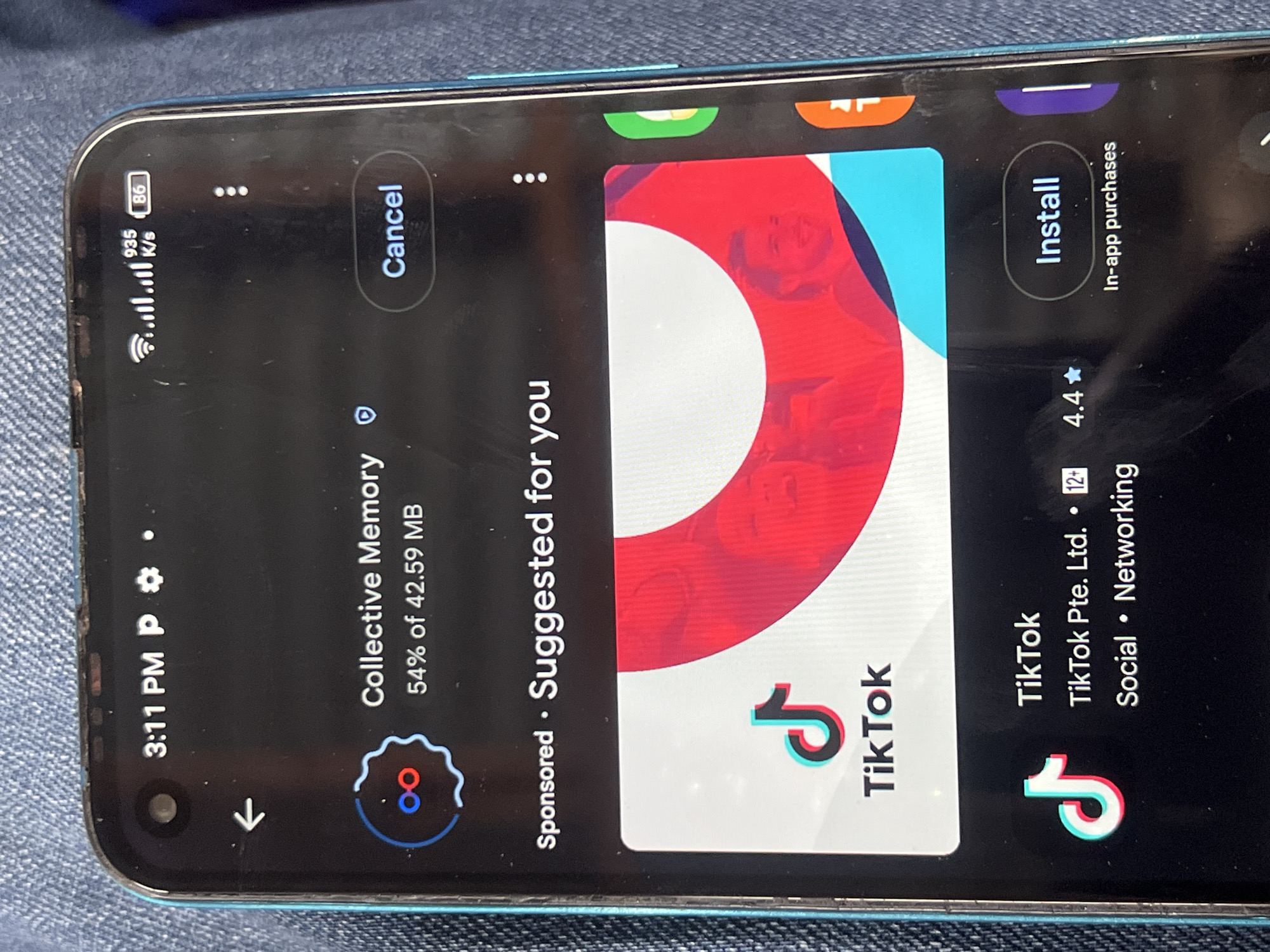 A close-up shot captures a smartphone screen displaying an app store interface, oriented in landscape mode, with the phone resting on a textured, denim-like surface. The screen shows a download in progress for an app called "Collective Memory," which is 54% complete out of 42.59 MB, with a "Cancel" button visible.
Below this, a sponsored suggestion for the "TikTok" app is featured, labeled "Sponsored. Suggested for you." TikTok is presented with its distinctive logo, a 4.4-star rating, and a 12+ age rating. It is identified as being from "TikTok Pte. Ltd." and categorized as "Social" and "Networking." An "Install" button is prominent, along with text indicating "In-app purchases" are available.
The phone's status bar at the top displays the time as "3:11 PM." The battery level is at 86%, and a strong Wi-Fi signal is present, showing a download speed of "935 K/s." The phone's screen has minor scratches, and the bezel shows some wear. This scene takes place in Jalingo, Nigeria.