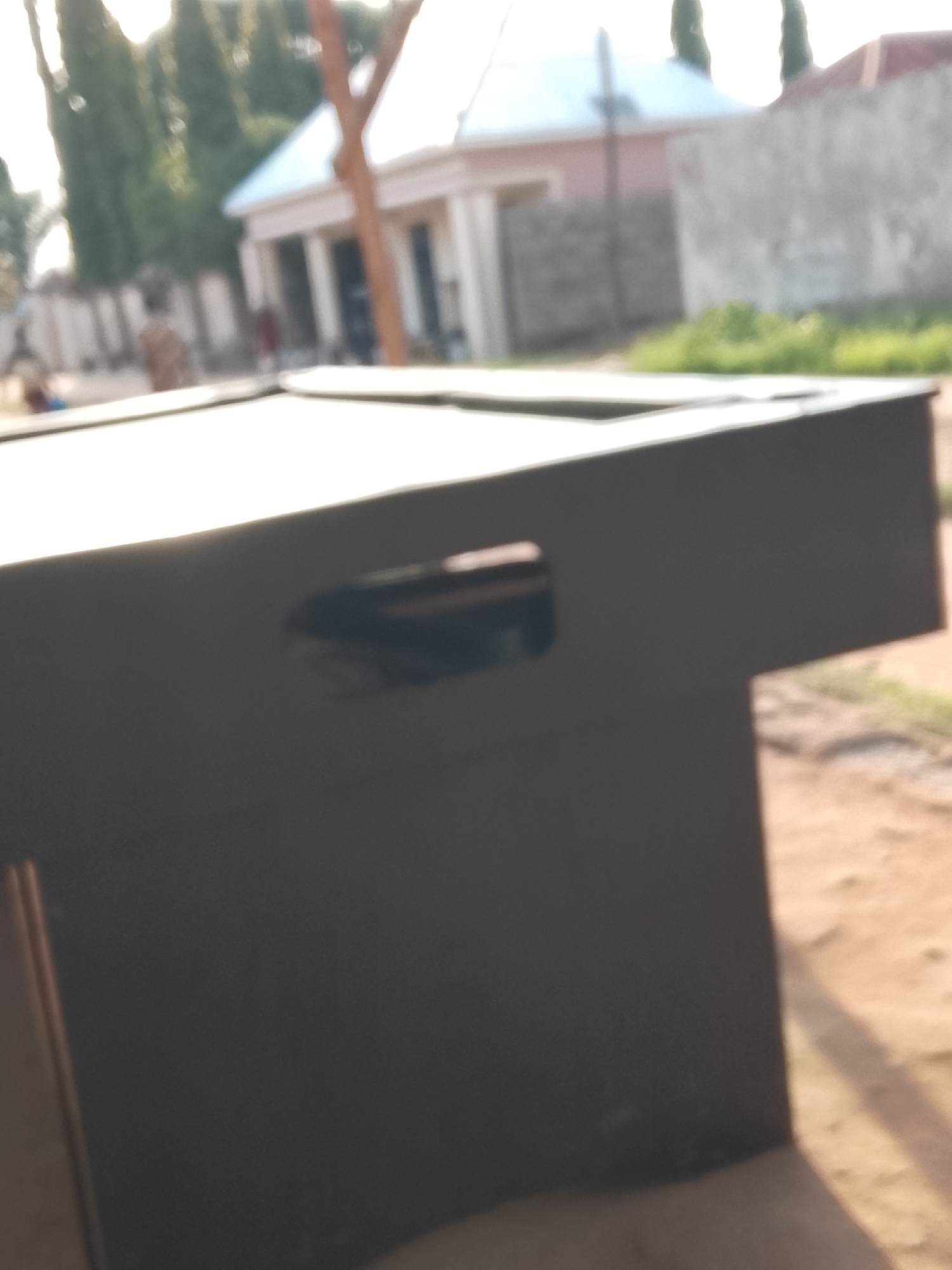 The image, taken in Jalingo, Nigeria, features a dark, rectangular object in the foreground, possibly a table or a chest, with a cutout handle on its side. The object is placed on sandy ground, with some dry grass and shadows visible. In the background, a blurred view of buildings and trees suggests an outdoor setting. A house with a light-colored roof and arched windows is visible, along with other structures and a wall. The lighting indicates it is likely daytime, possibly late afternoon due to the shadows. No people are clearly discernible in the scene.