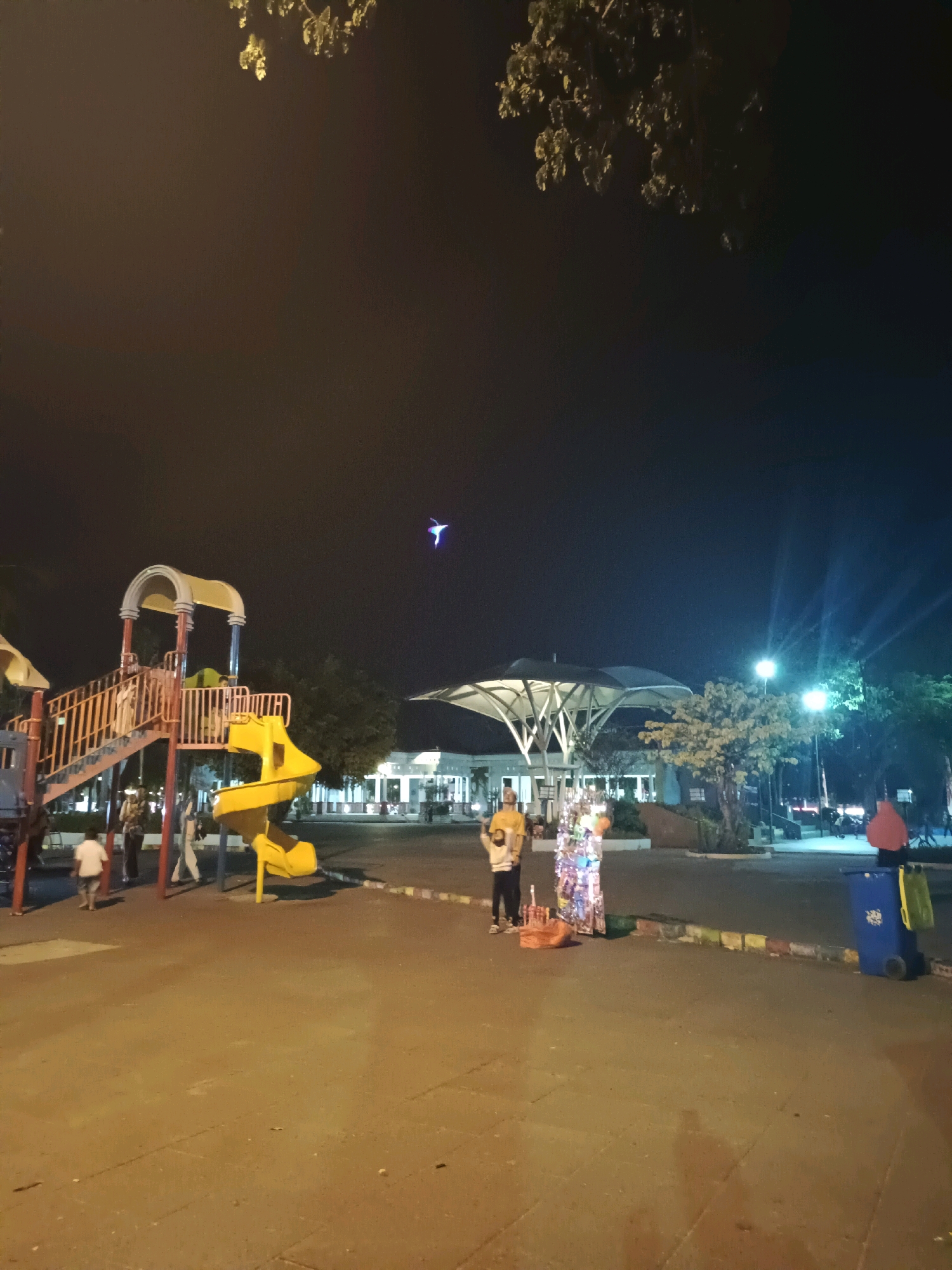 The image depicts an outdoor scene at night, likely in a public park or recreational area, possibly in Lamongan, Indonesia, given the context. In the foreground, there is a children's playground with a yellow slide and red and yellow structures. Several people are present, including children near the playground and two individuals standing near a stall with colorful items, possibly vendors. A blue trash can with a green bag is visible to the right. In the background, there are buildings with bright lighting and trees. The sky is dark, with a small, brightly lit object, possibly a drone or a kite, visible in the upper center of the frame. The overall lighting suggests it is nighttime, with artificial lights illuminating the scene.