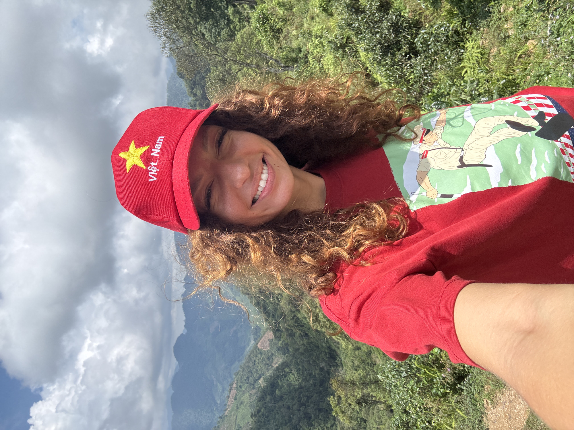 A young woman with curly brown hair and a red baseball cap featuring a gold star and the words "Việt Nam" is taking a selfie. She is smiling, and the background shows a mountainous landscape with vegetation. The sky is cloudy. The woman is wearing a red t-shirt with a graphic design. The selfie appears to be taken in a natural environment, consistent with the mountainous area of Suối Giàng, Vietnam.