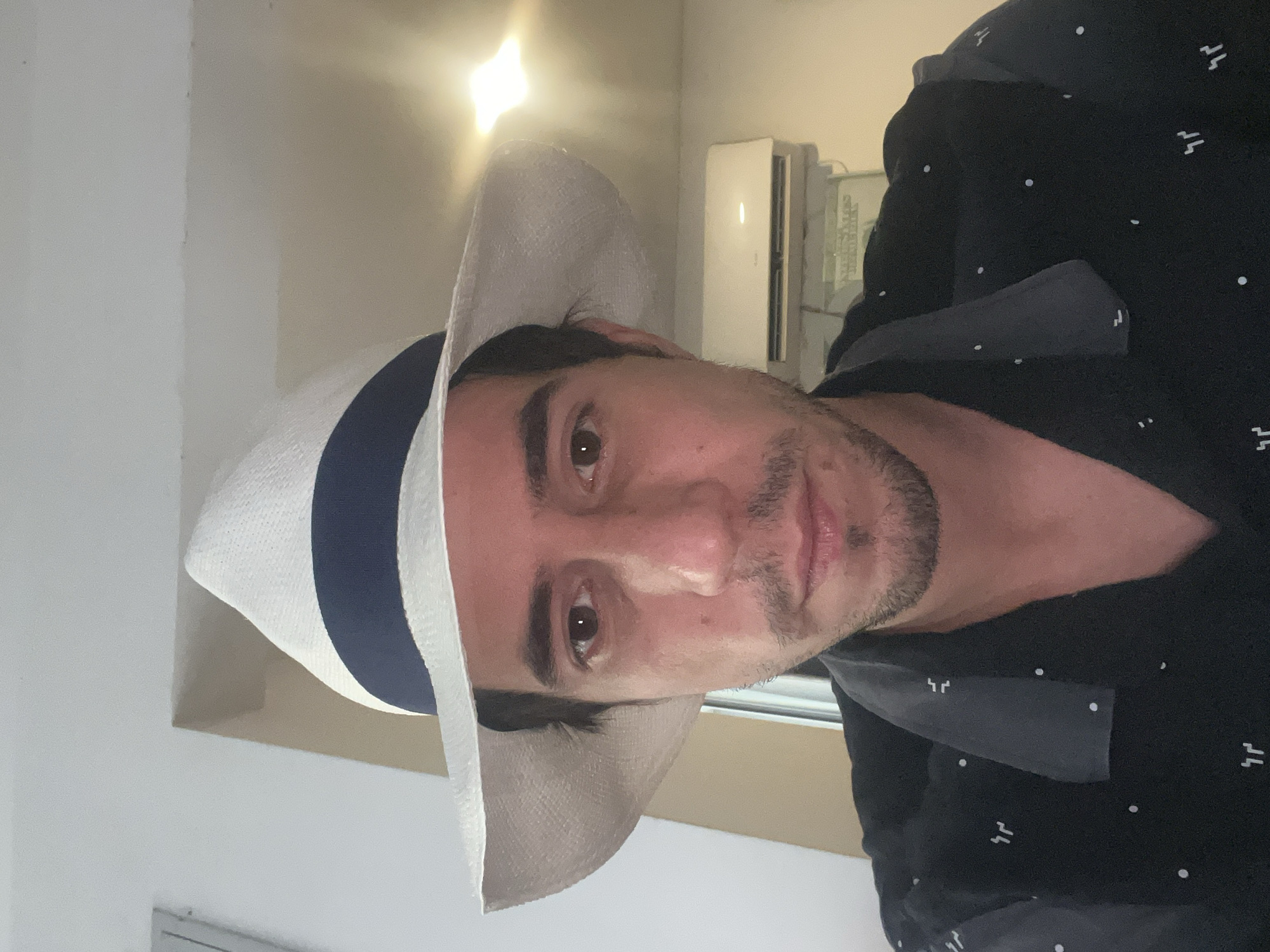 A man is indoors, wearing a white hat with a navy blue band and a black button-down shirt. The location appears to be a generic room, possibly in Buenos Aires, Argentina, but there is no specific evidence to confirm the city. An air conditioning unit is visible on the wall behind him. The man is looking directly at the camera.