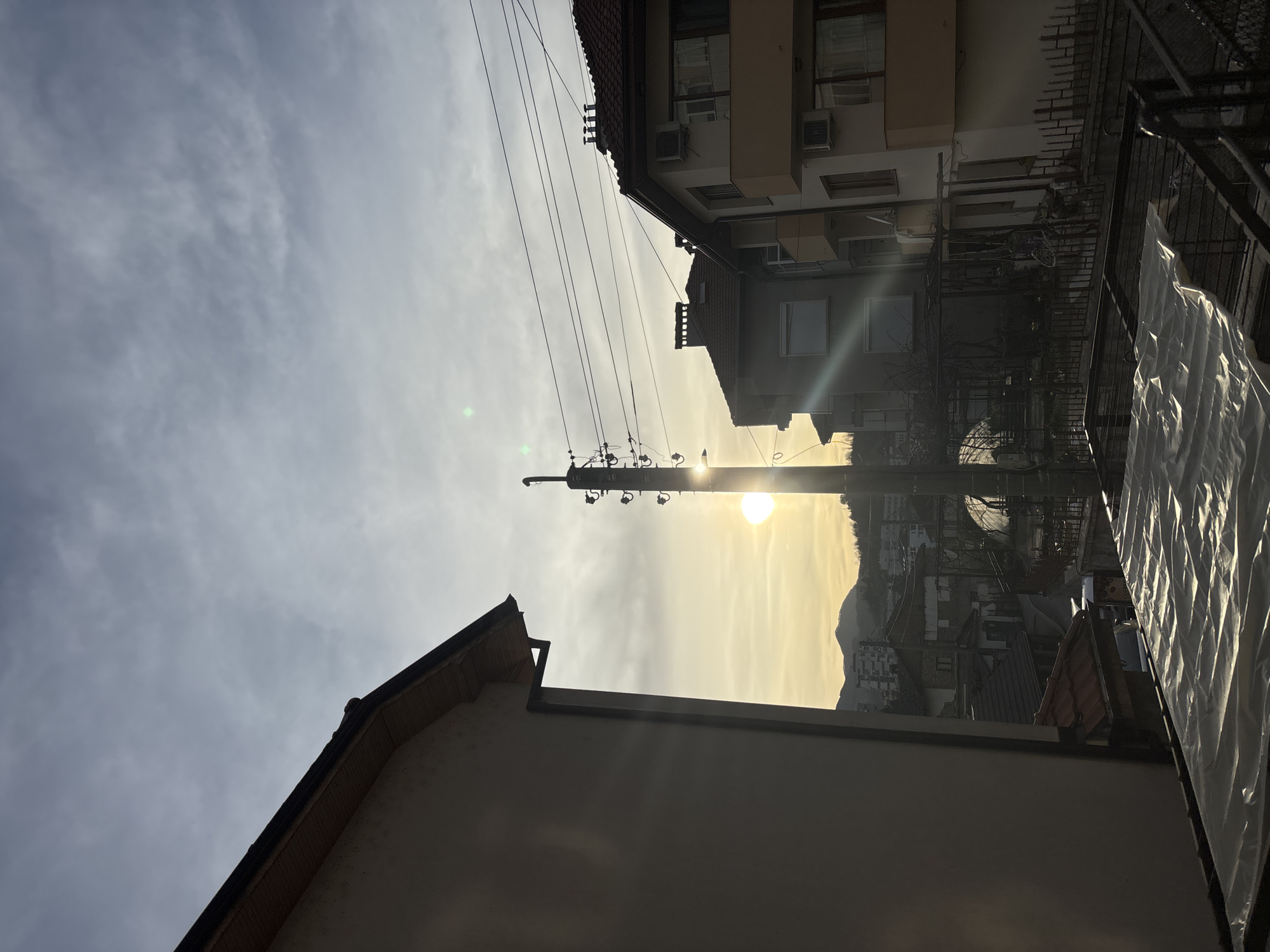 The image, taken in Zlatograd, Bulgaria, shows a view of buildings and a utility pole silhouetted against a bright sky with the sun partially obscured by clouds. In the foreground, the corner of a light-colored building extends upwards, with a roofline visible. The sun is centered and positioned behind the utility pole. Power lines stretch across the sky. Beyond the utility pole, apartment buildings are visible. In the right foreground, there is a fence, and a transparent plastic sheet is partially visible. In the background, a hill. No people are visible.